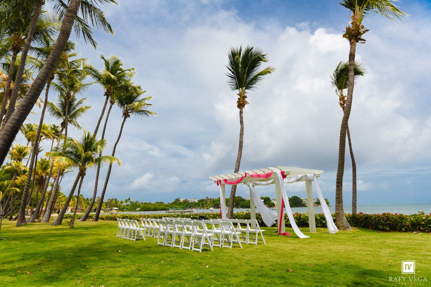 Outdoor beach wedding setup with white chairs, floral arch, palm trees, and green lawn under a dramatic cloudy sky.
