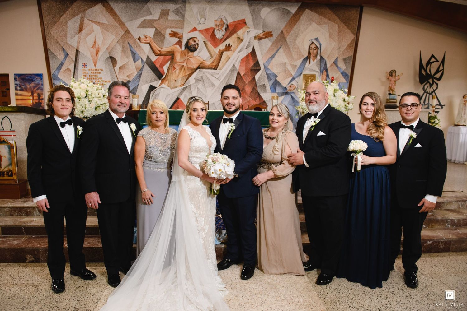 Wedding party posing in formal attire before a religious mural, bride in white gown holding bouquet with groom in navy suit.