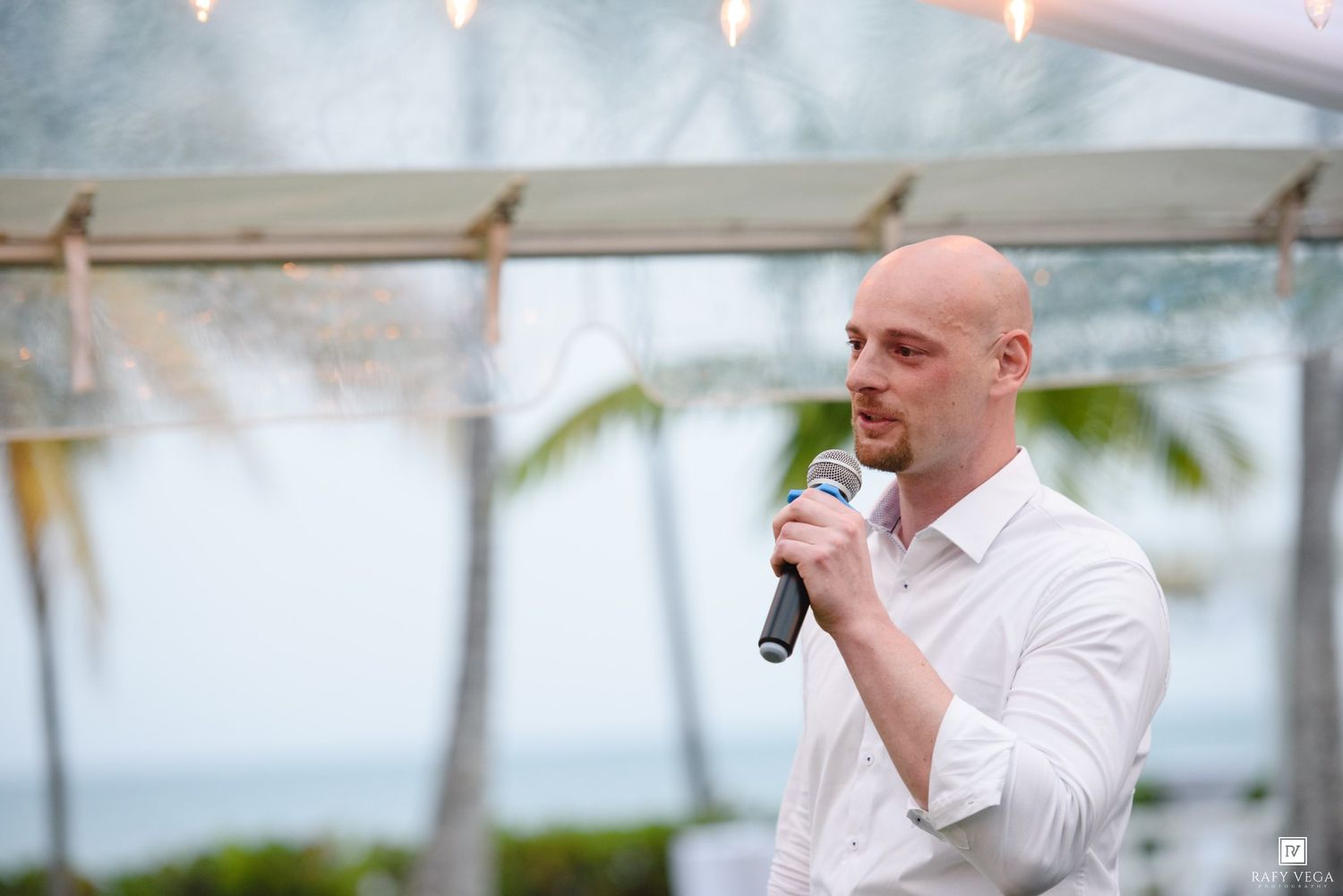 Bald man in white shirt speaking into microphone at outdoor tropical event with palm trees in background.