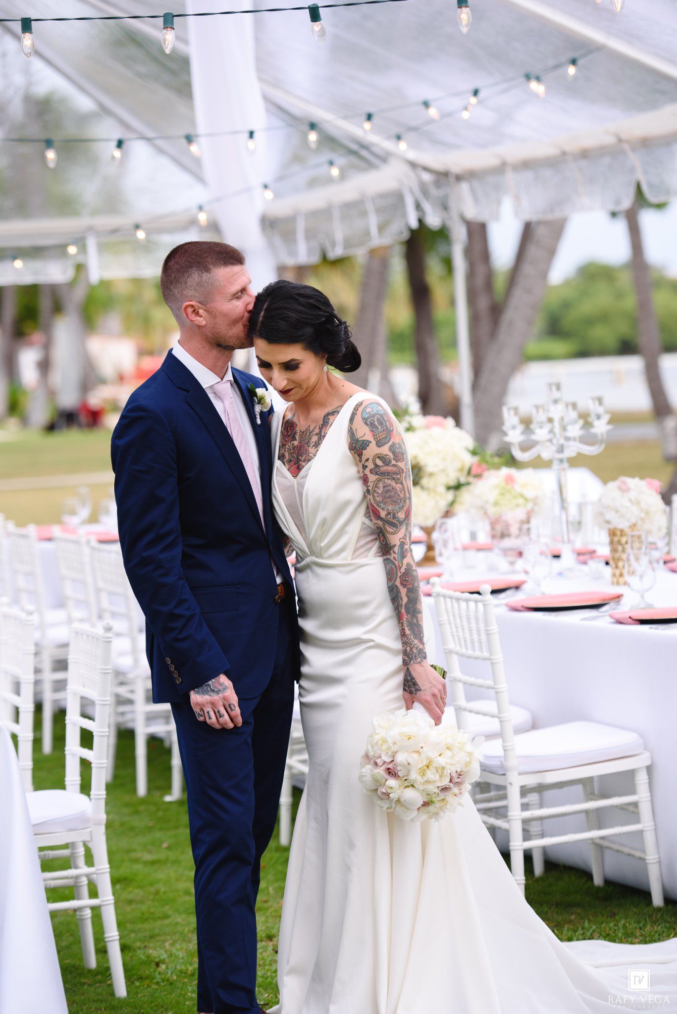 Bride and groom share a tender moment at elegant outdoor wedding reception with white chairs and floral centerpieces.