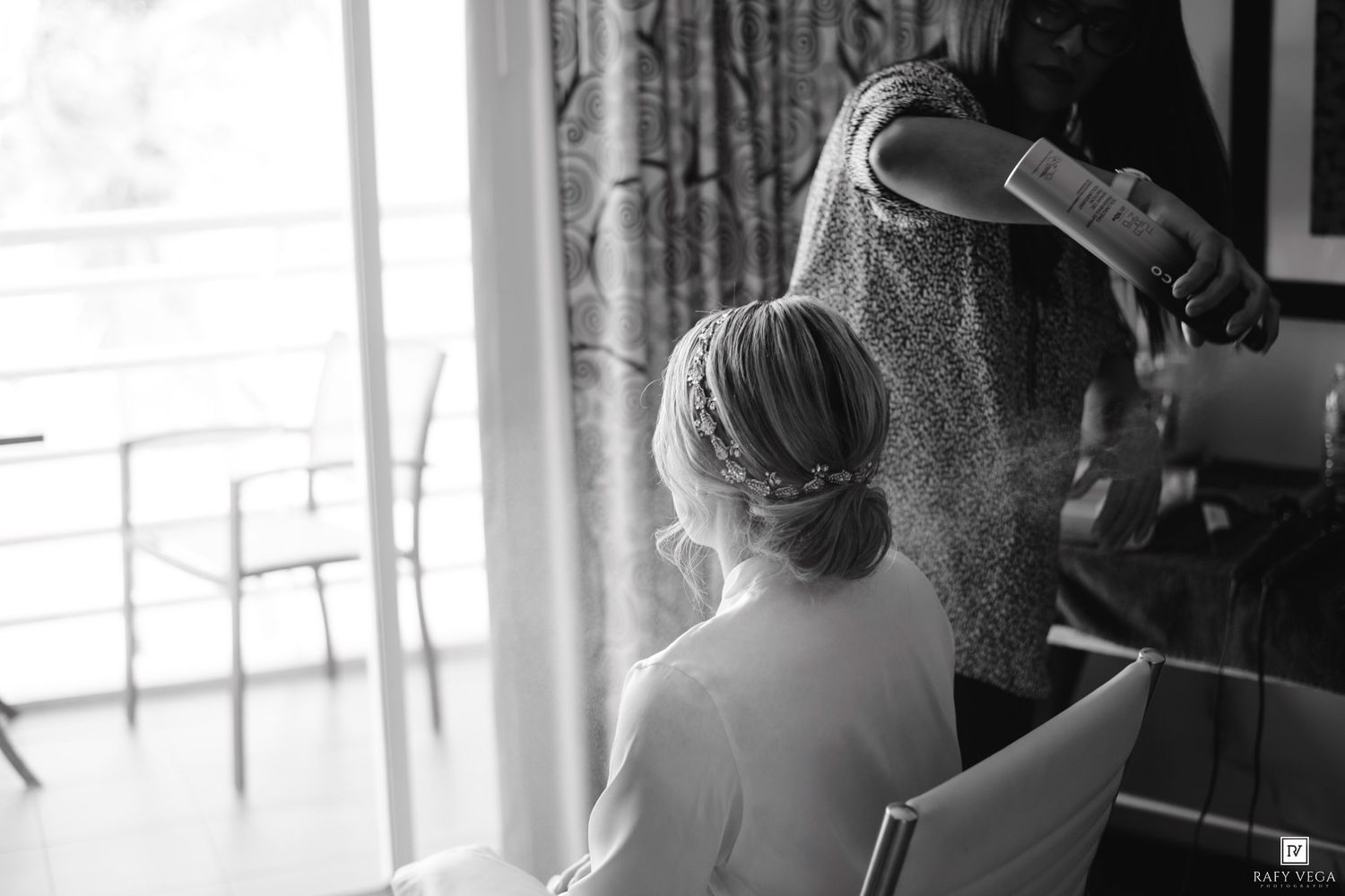 Hairstylist applying hairspray to a seated bride with an elegant updo during wedding preparations in black and white.