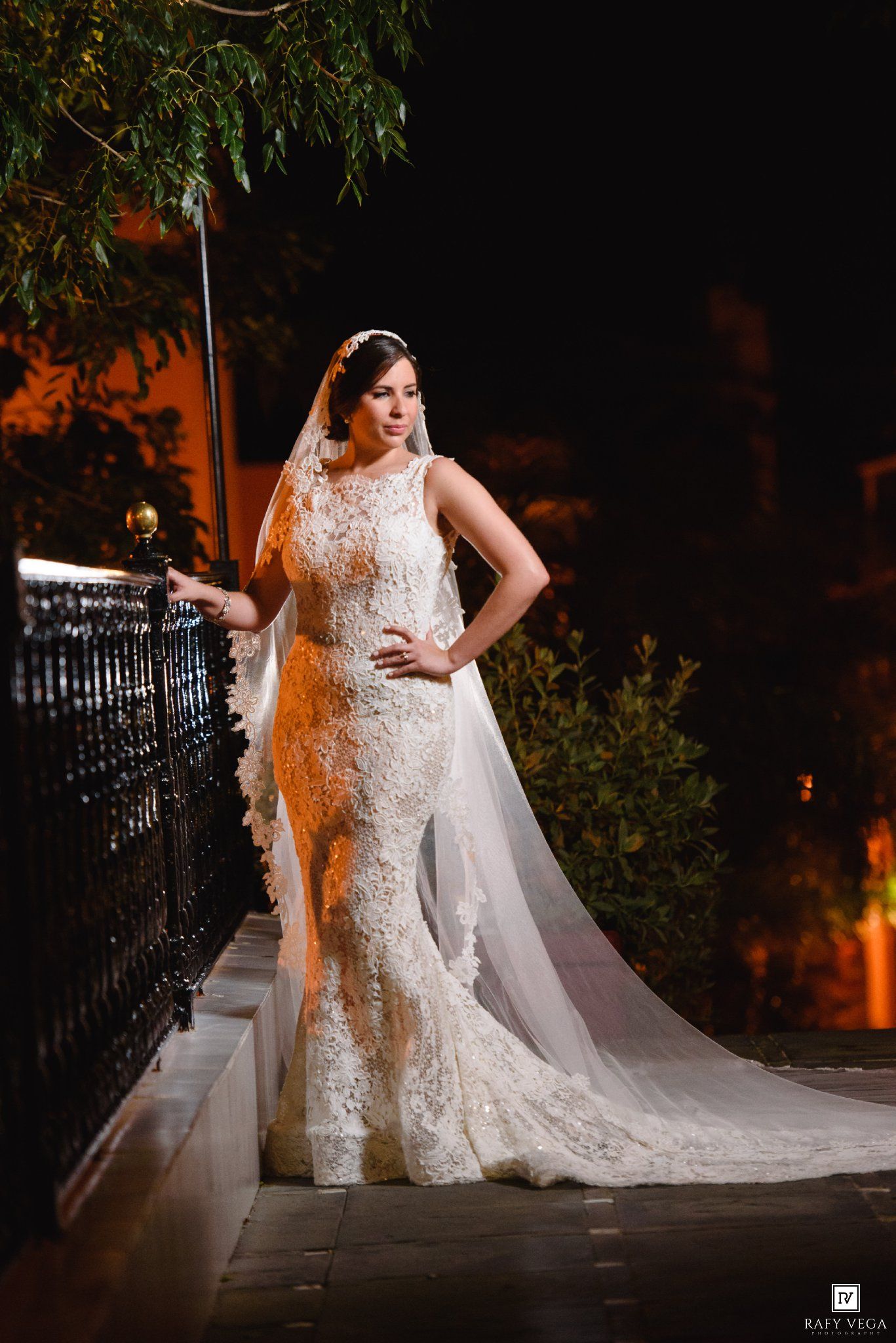 Bride in elegant lace mermaid wedding gown with cathedral veil posing on outdoor staircase at night.