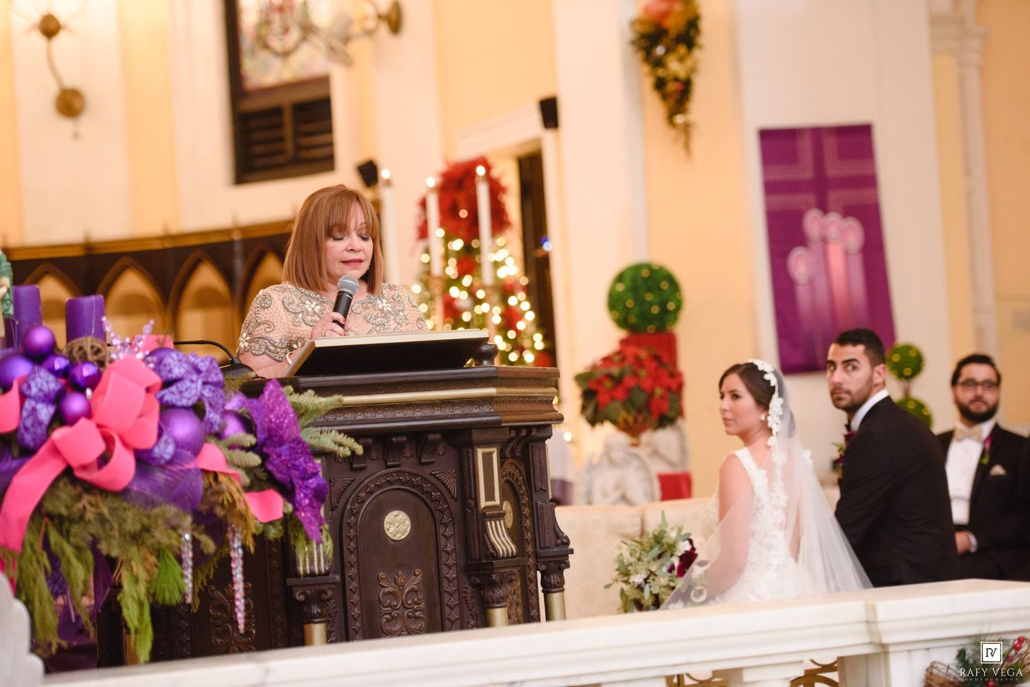 A woman speaks at a podium during a church wedding ceremony with a bride and groom standing nearby.