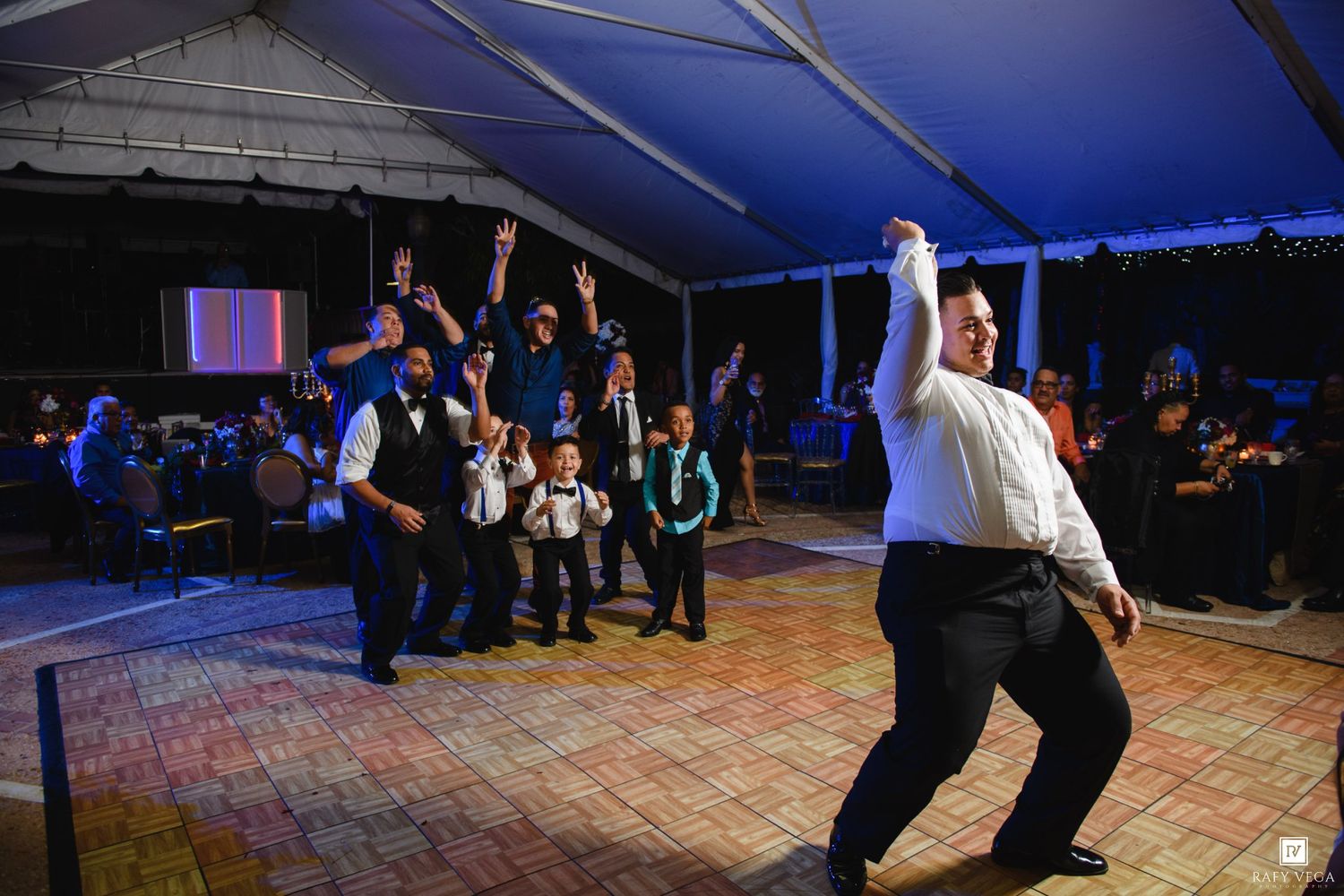 Groom tosses garter to groomsmen on dance floor at outdoor tented wedding reception at night.