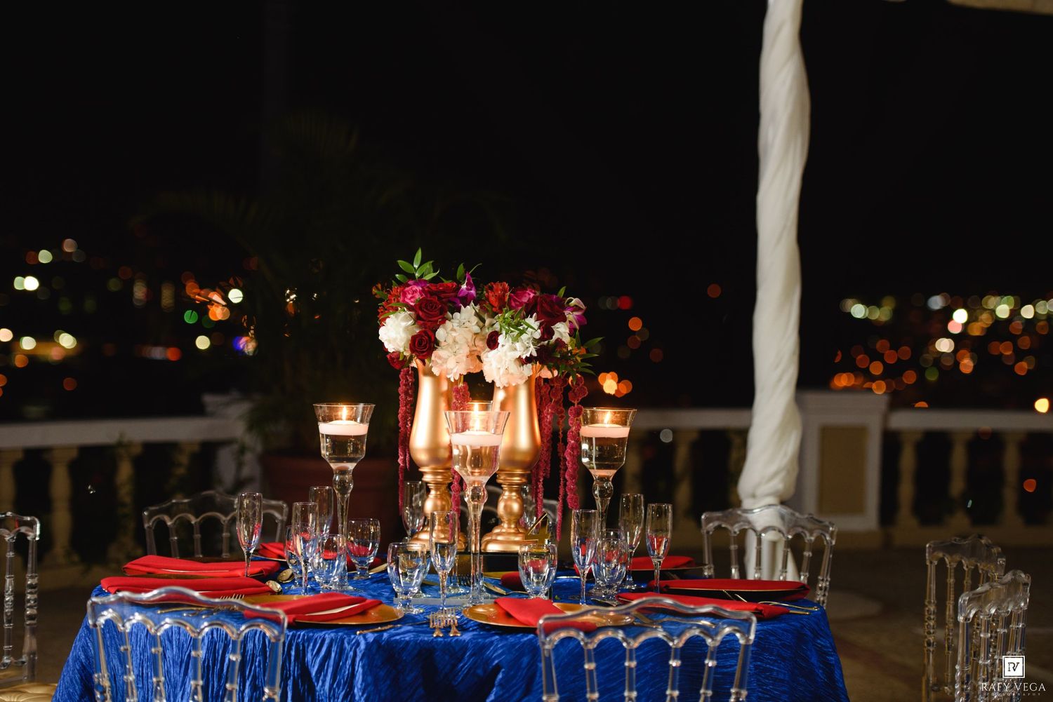 Elegant outdoor wedding reception table with blue velvet linen, red napkins, gold candelabras, and floral centerpiece at night.