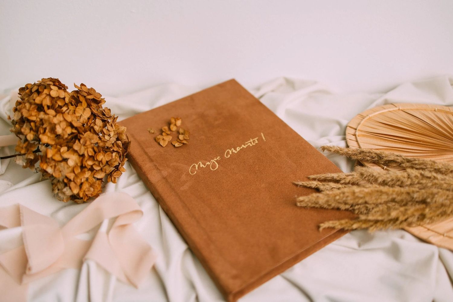Brown velvet guest book with cursive text, surrounded by dried flowers and wheat stems on white fabric.