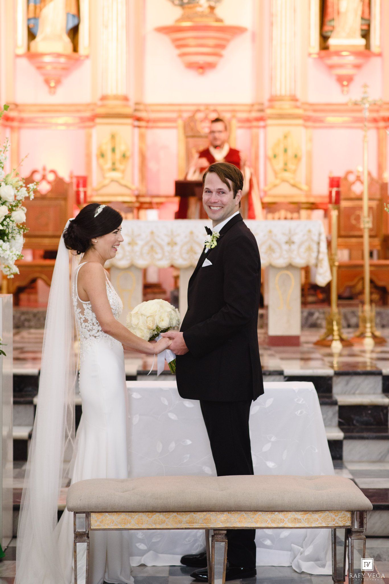 Bride and groom holding hands at church altar during wedding ceremony, bride in white gown with veil holding bouquet.
