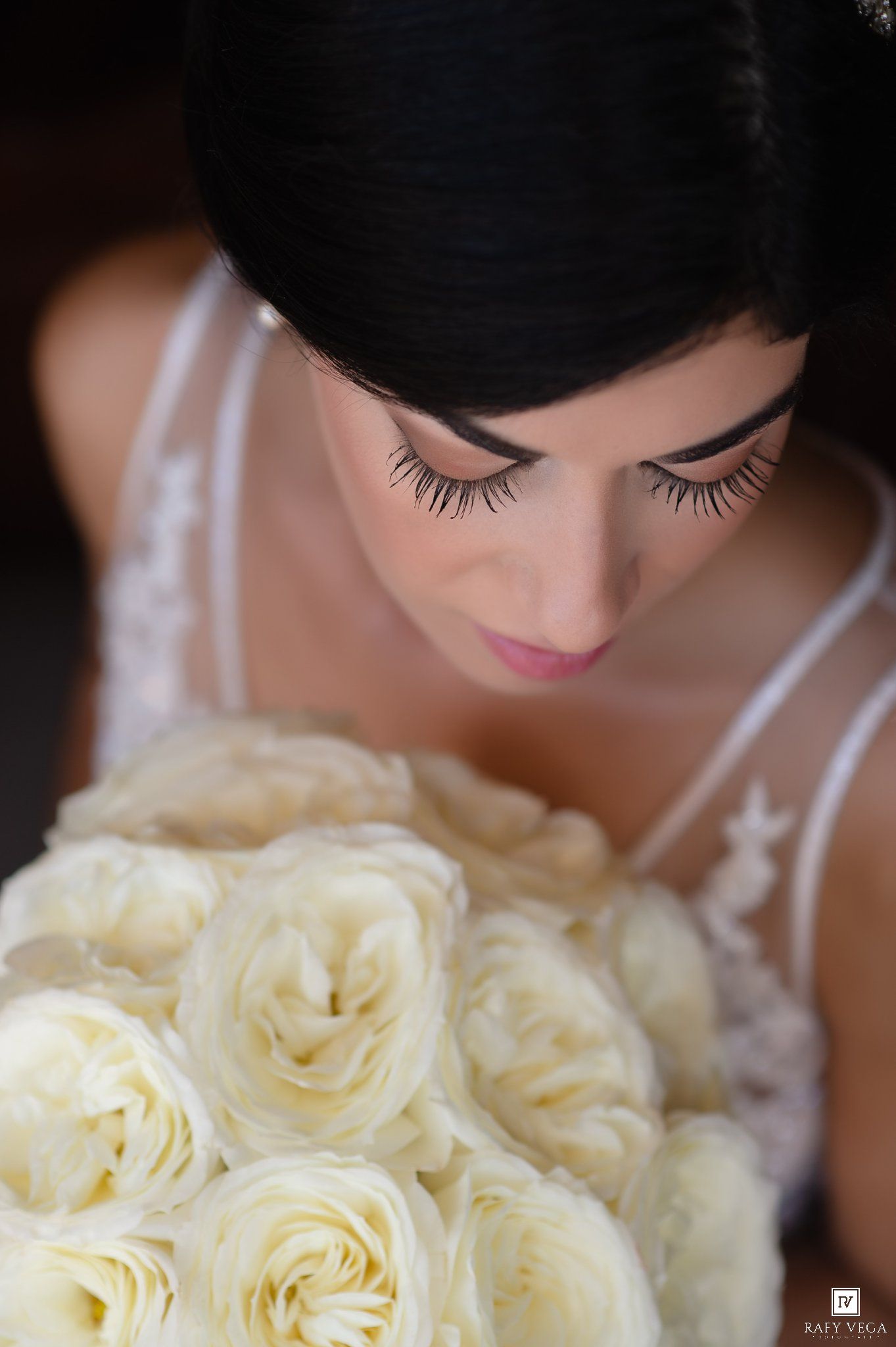 Bride with dark hair and dramatic lashes looks down at a bouquet of white roses in a wedding gown.