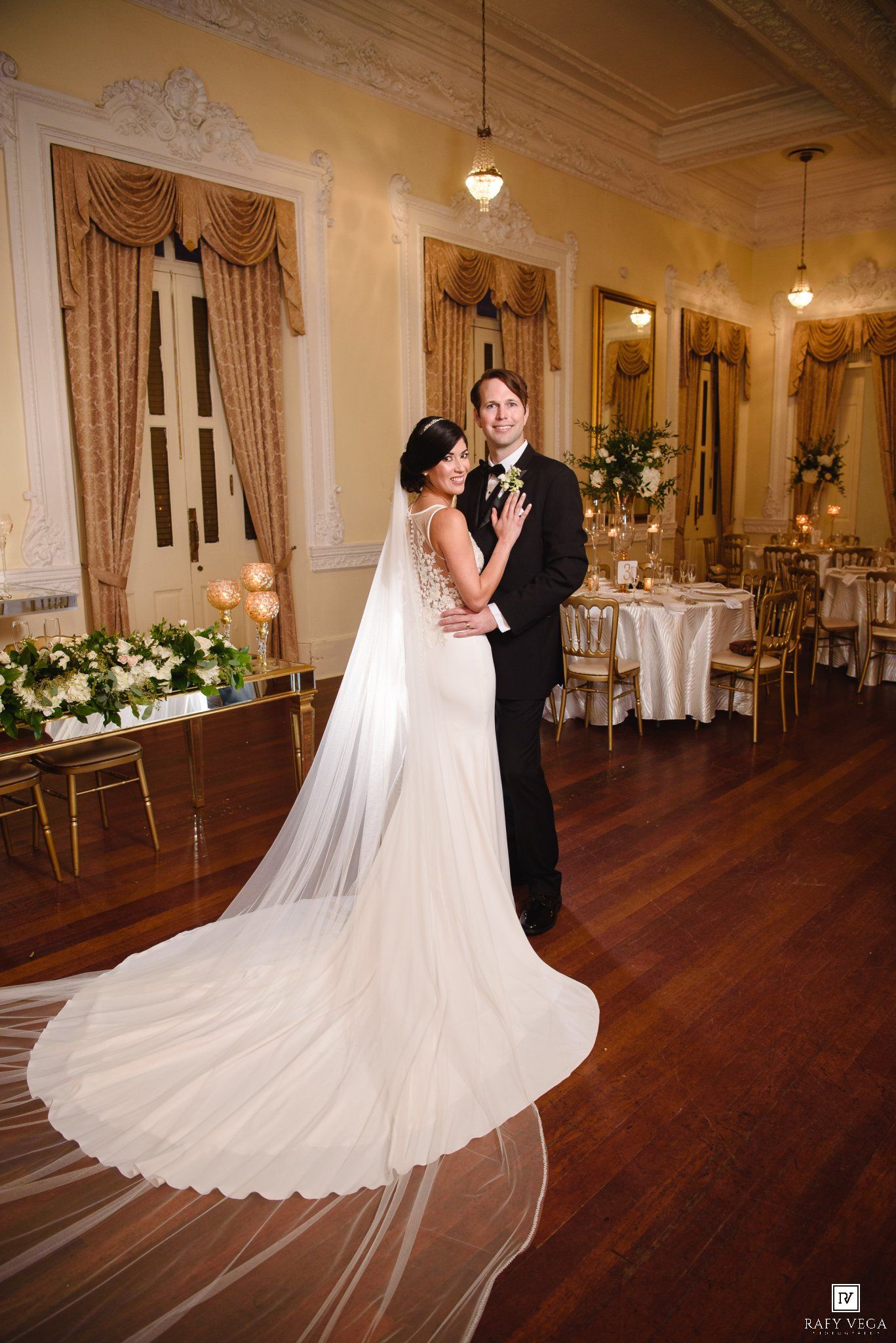 Bride in white gown with long veil and groom in tuxedo embrace on dance floor of elegant ballroom.
