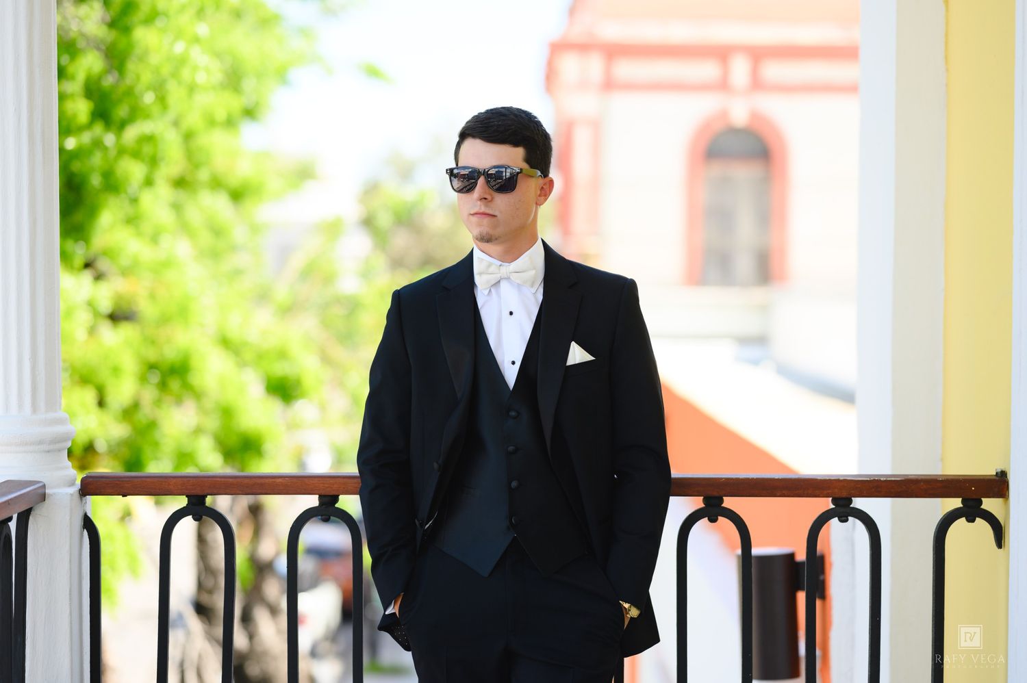 Stylish groom in black tuxedo and sunglasses poses on balcony outdoors on sunny wedding day.