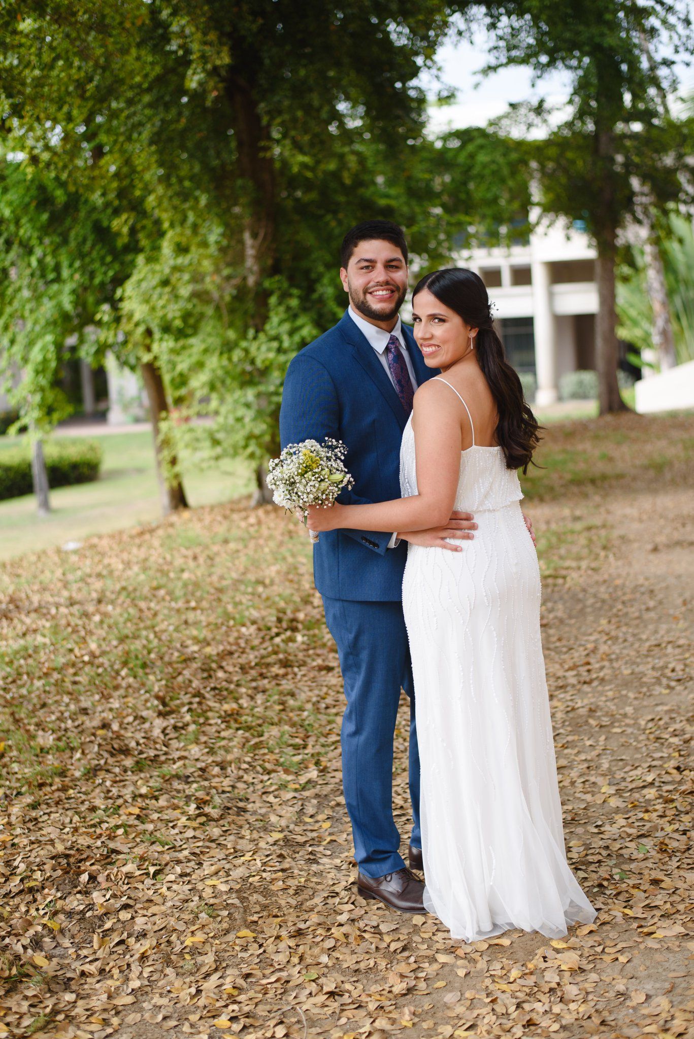 Smiling bride and groom pose outdoors in a park, she in white gown holding bouquet, he in navy suit.