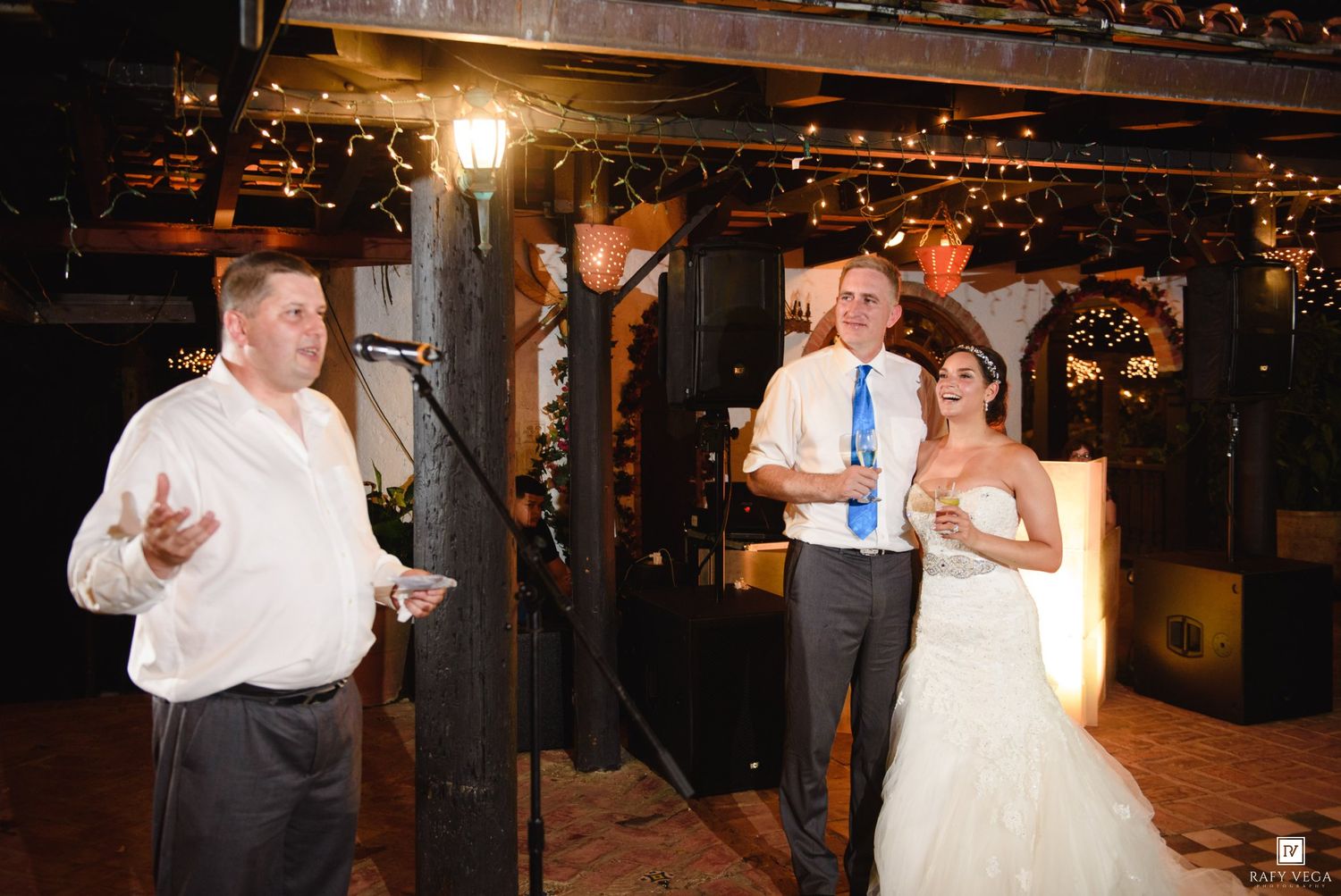 A groom and bride stand together at their wedding reception while a guest delivers a heartfelt speech indoors.