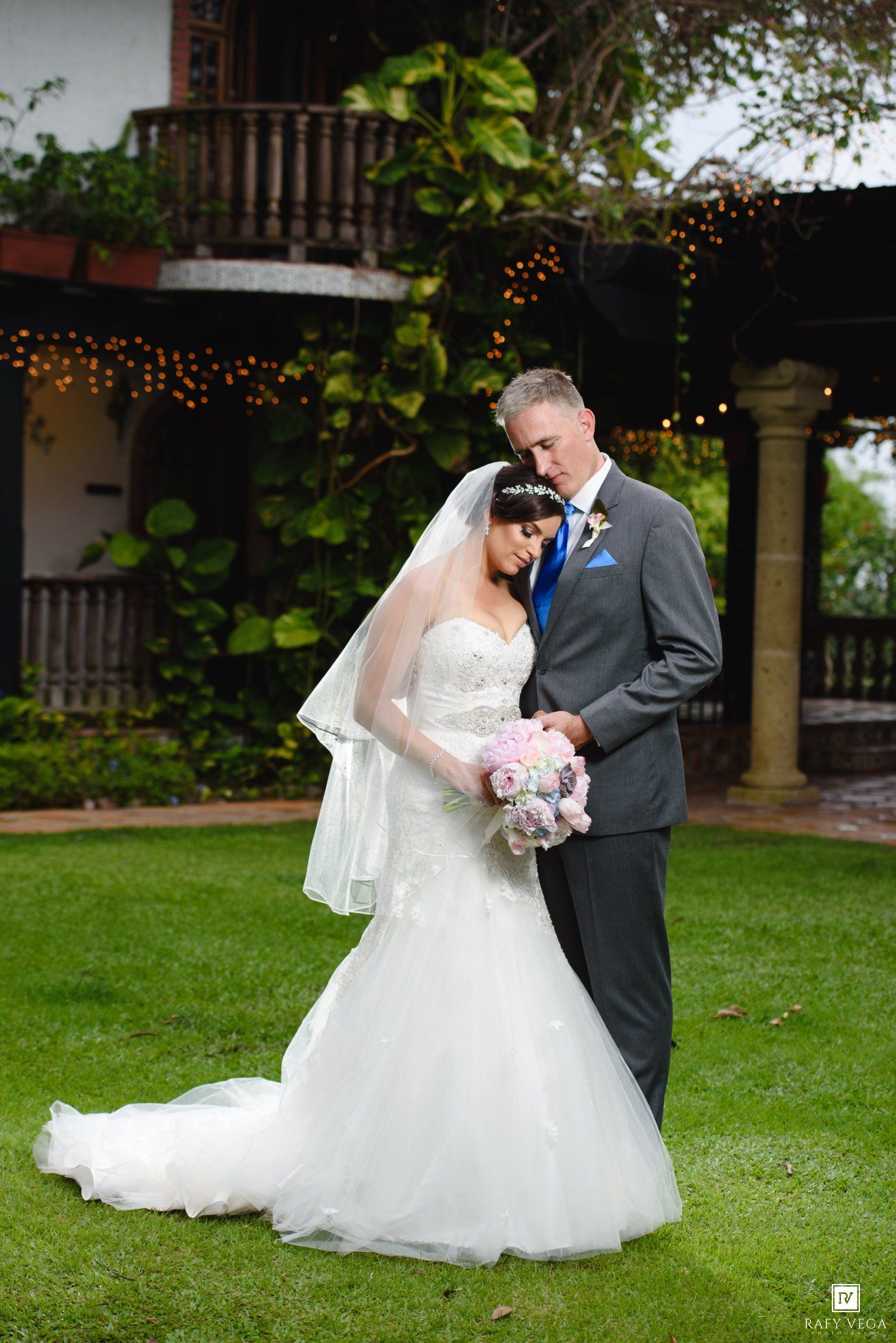 Bride in white gown with veil holds pink bouquet as groom in gray suit embraces her on green lawn.