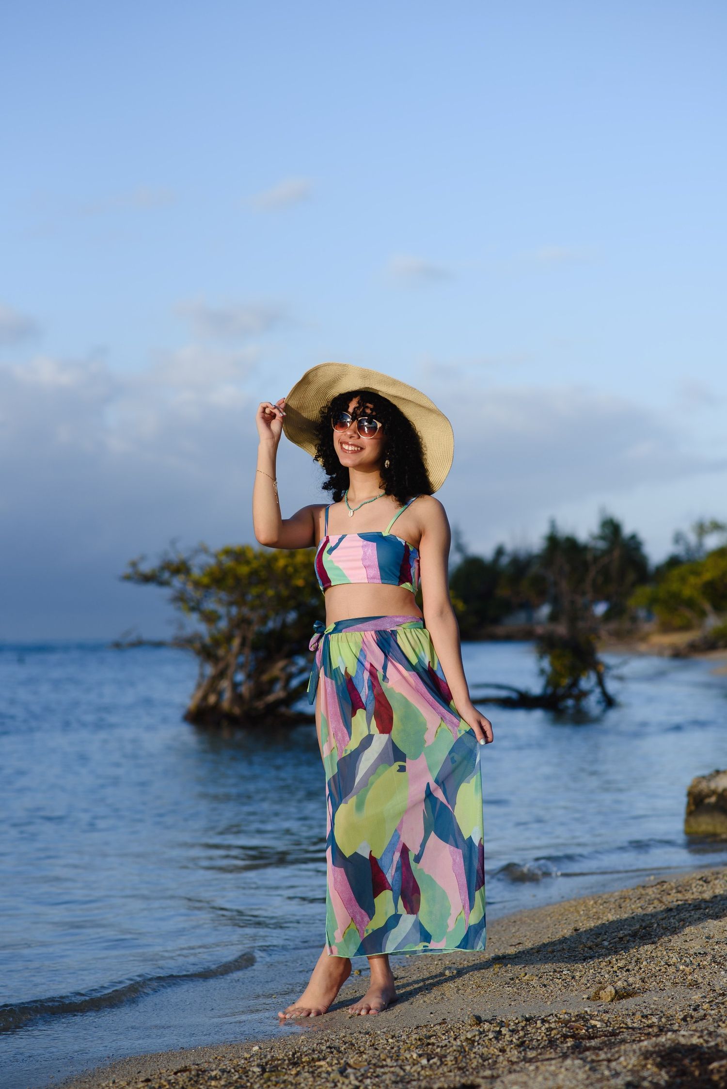 Woman in colorful crop top, maxi skirt, and sun hat poses on a tropical beach at sunset.