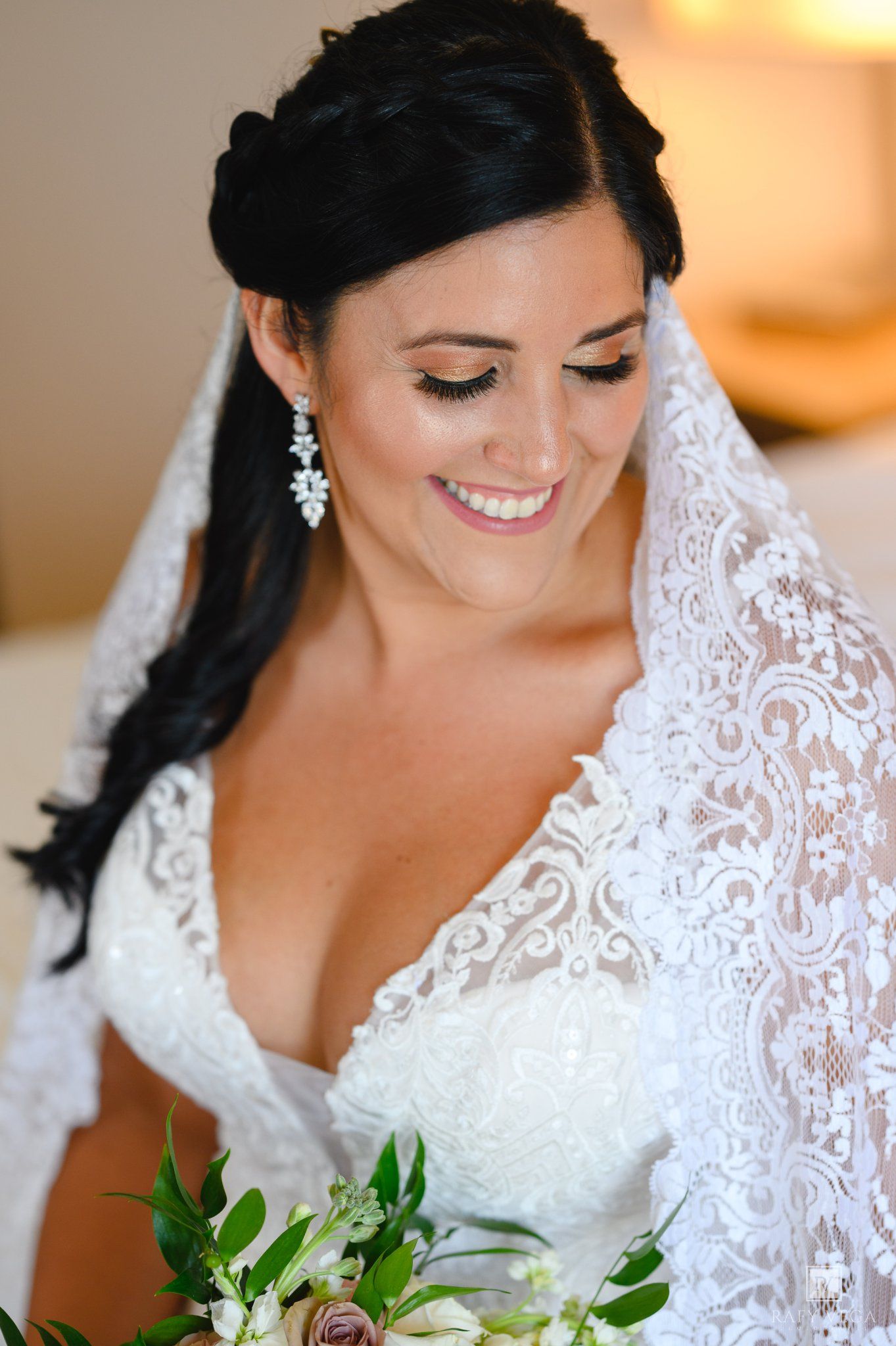 Smiling bride in lace wedding dress and veil holds bouquet, wearing crystal earrings with elegant updo hairstyle.