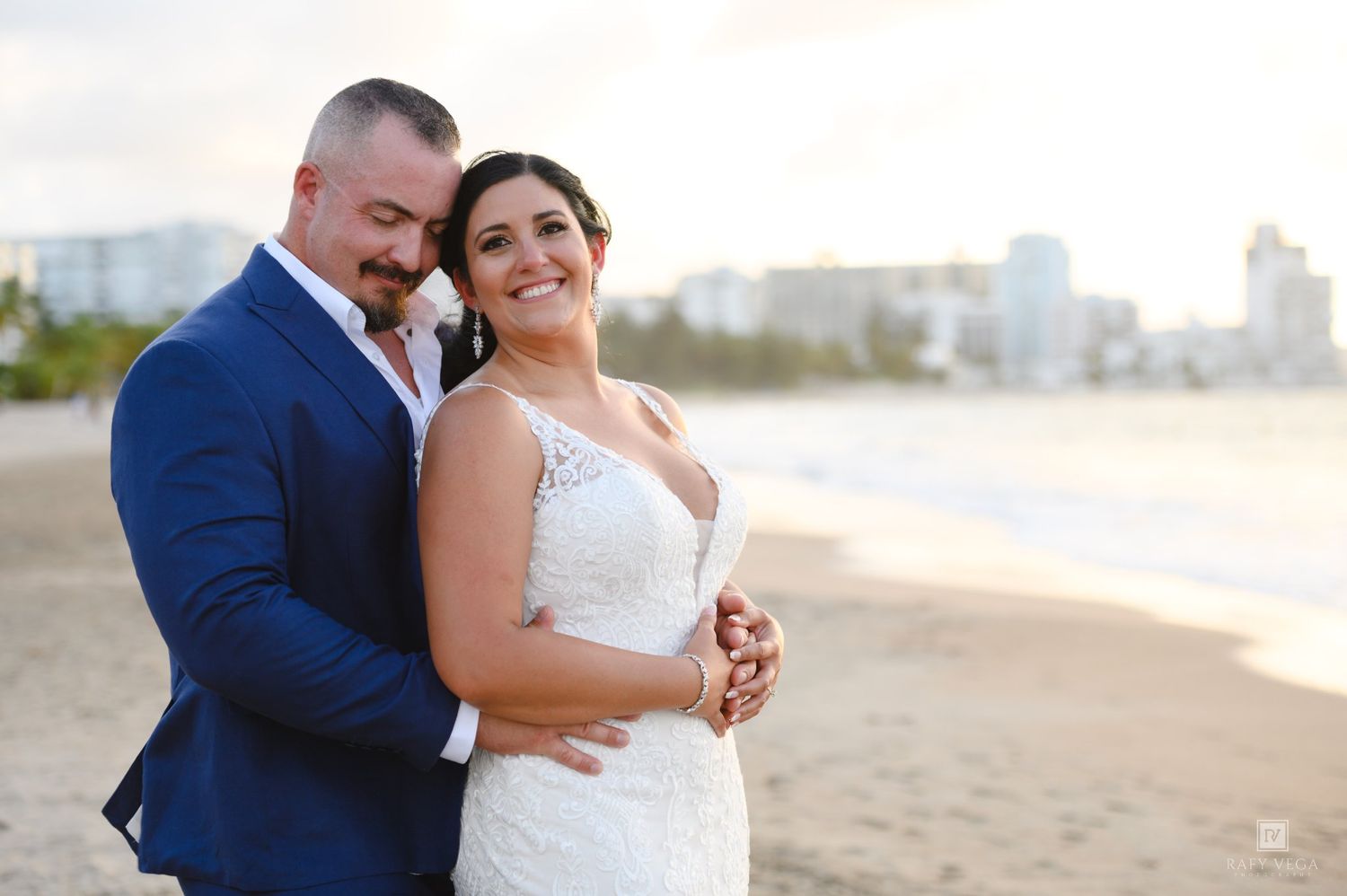 Bride and groom embracing on a beach at sunset, wearing a white lace gown and blue suit.
