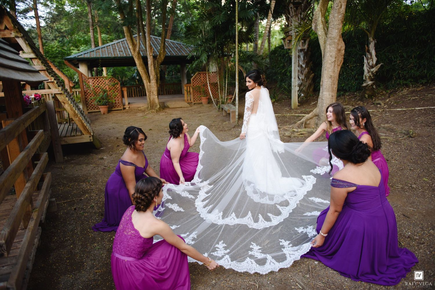 Bride in white gown with long lace veil held by bridesmaids in purple dresses at outdoor wedding venue.
