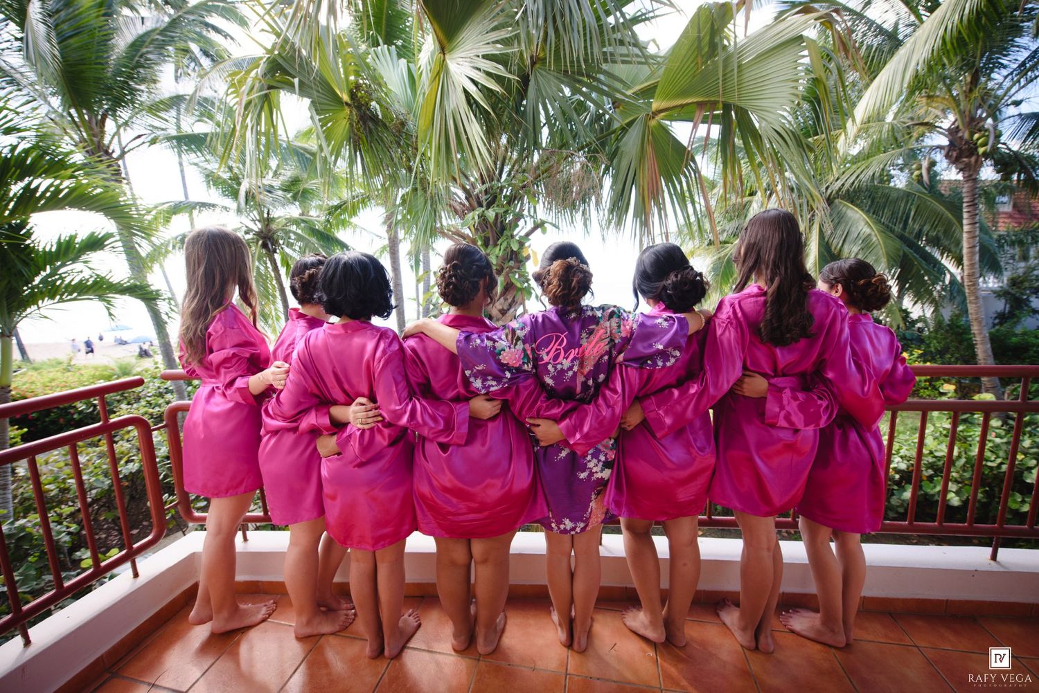 Bridesmaids in matching pink robes and bride in floral kimono stand on tropical resort balcony.