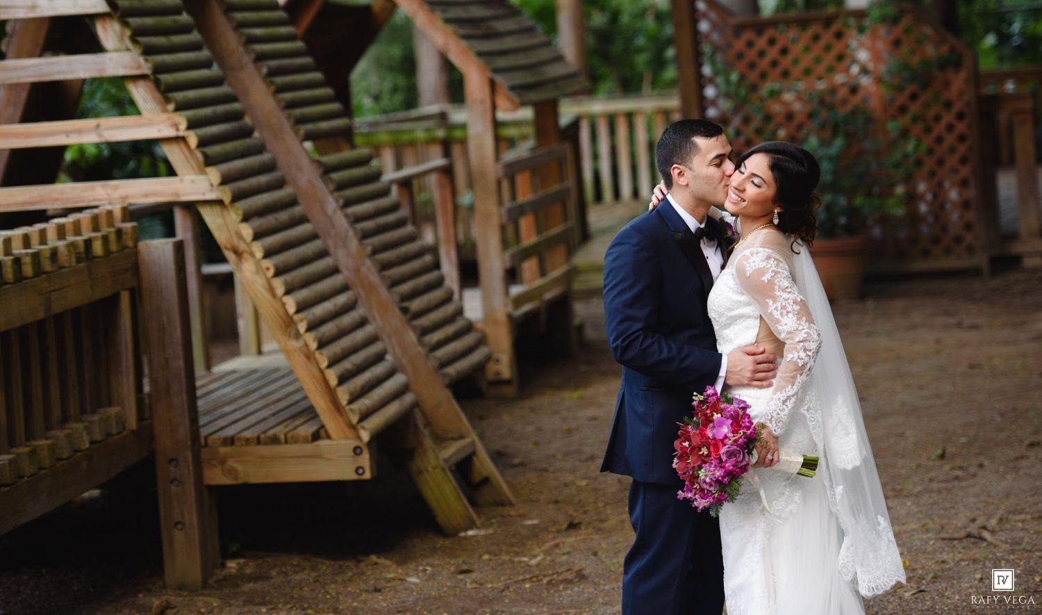 A bride and groom share a romantic kiss outdoors near wooden stairs, with the bride holding a vibrant pink bouquet.