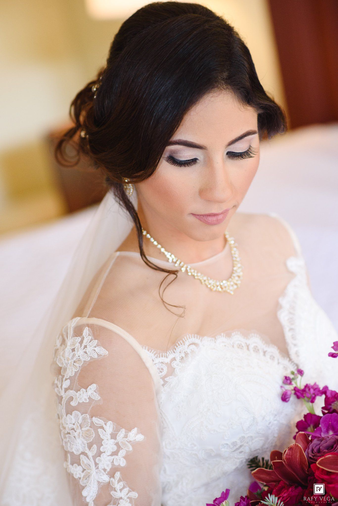 Bride in lace wedding dress and pearl necklace holding purple bouquet with elegant updo and bridal makeup.