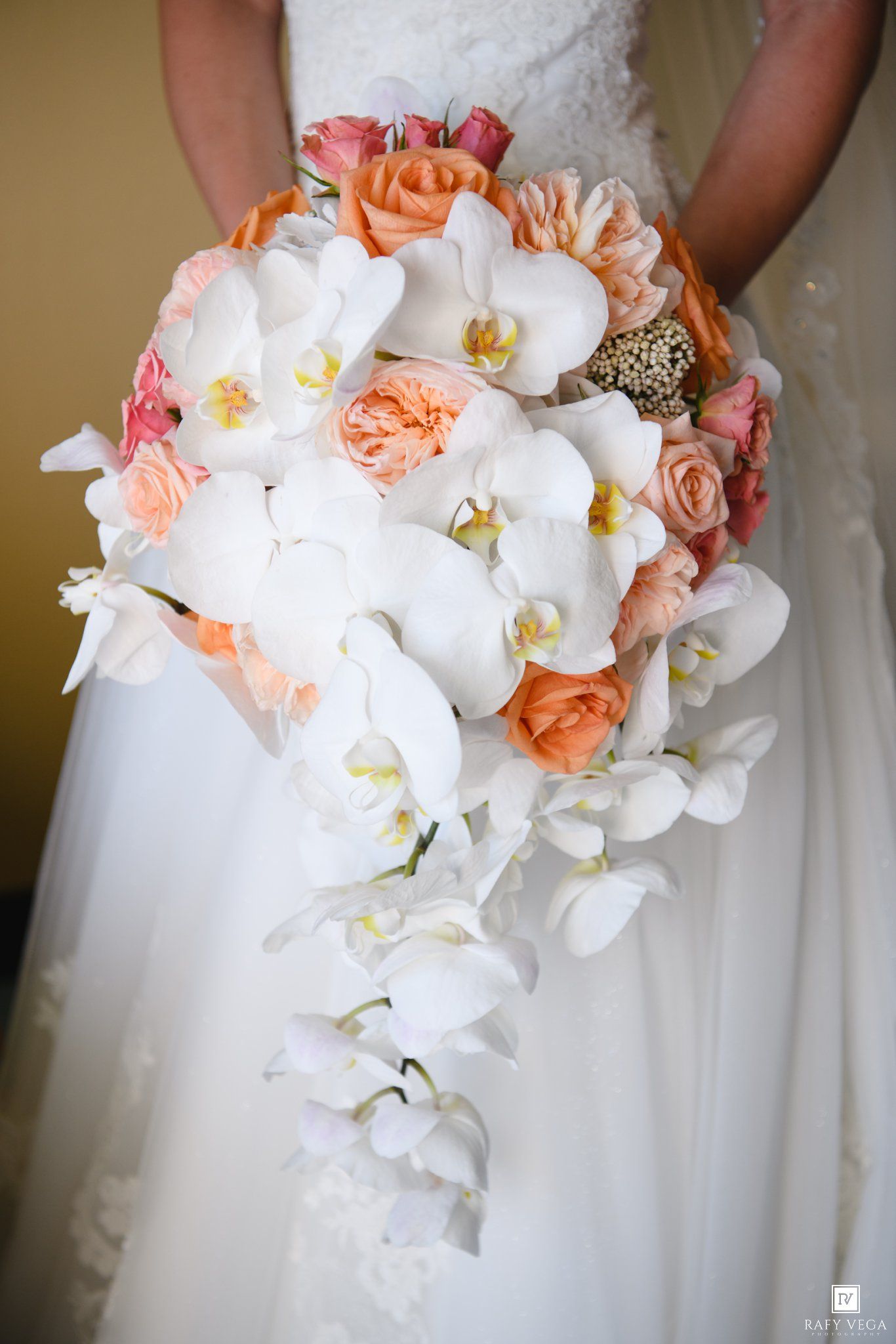 Bridal cascading bouquet with white phalaenopsis orchids and peach garden roses held by bride in white gown.