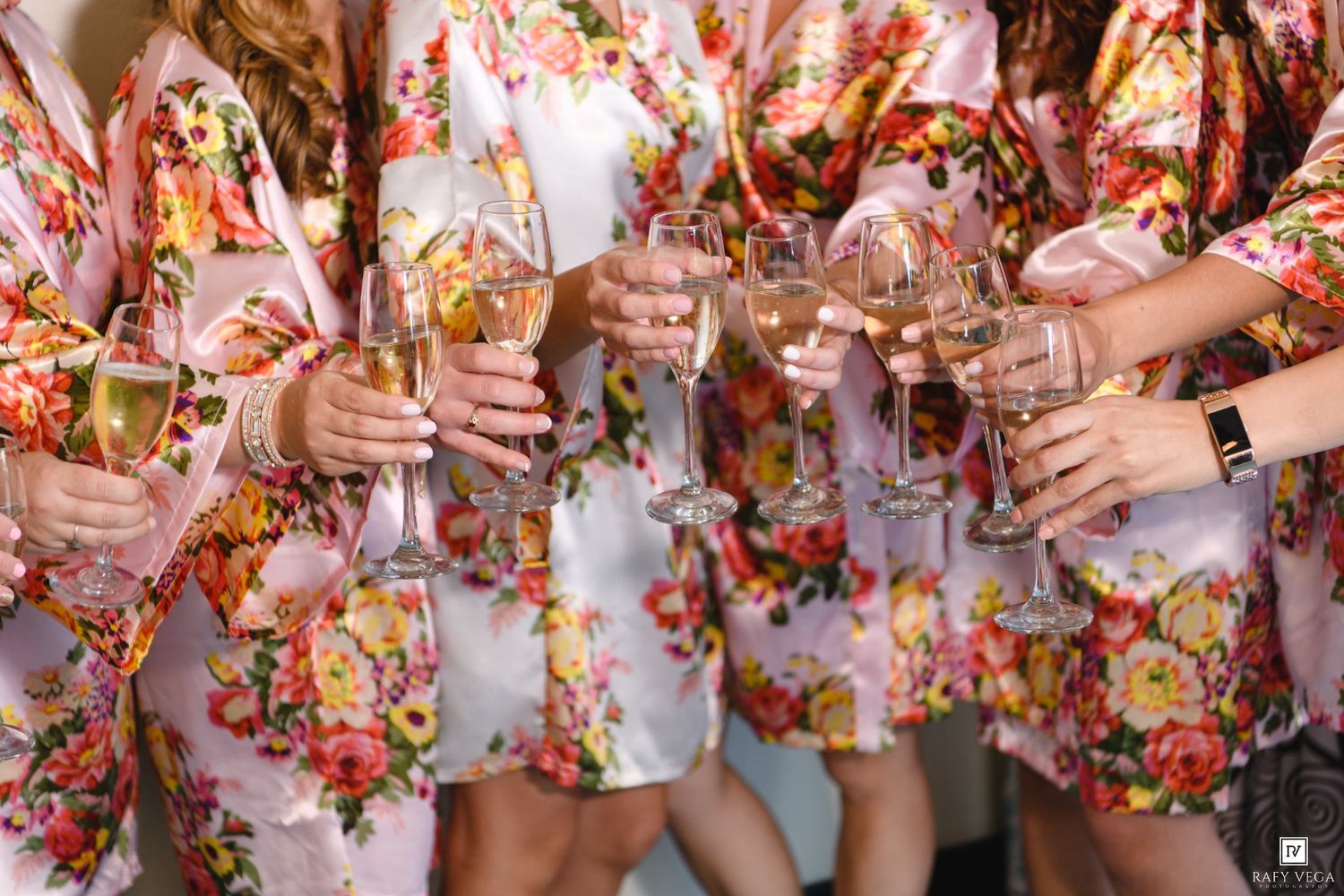 Bridesmaids in matching floral satin robes toasting with champagne glasses during wedding preparations.