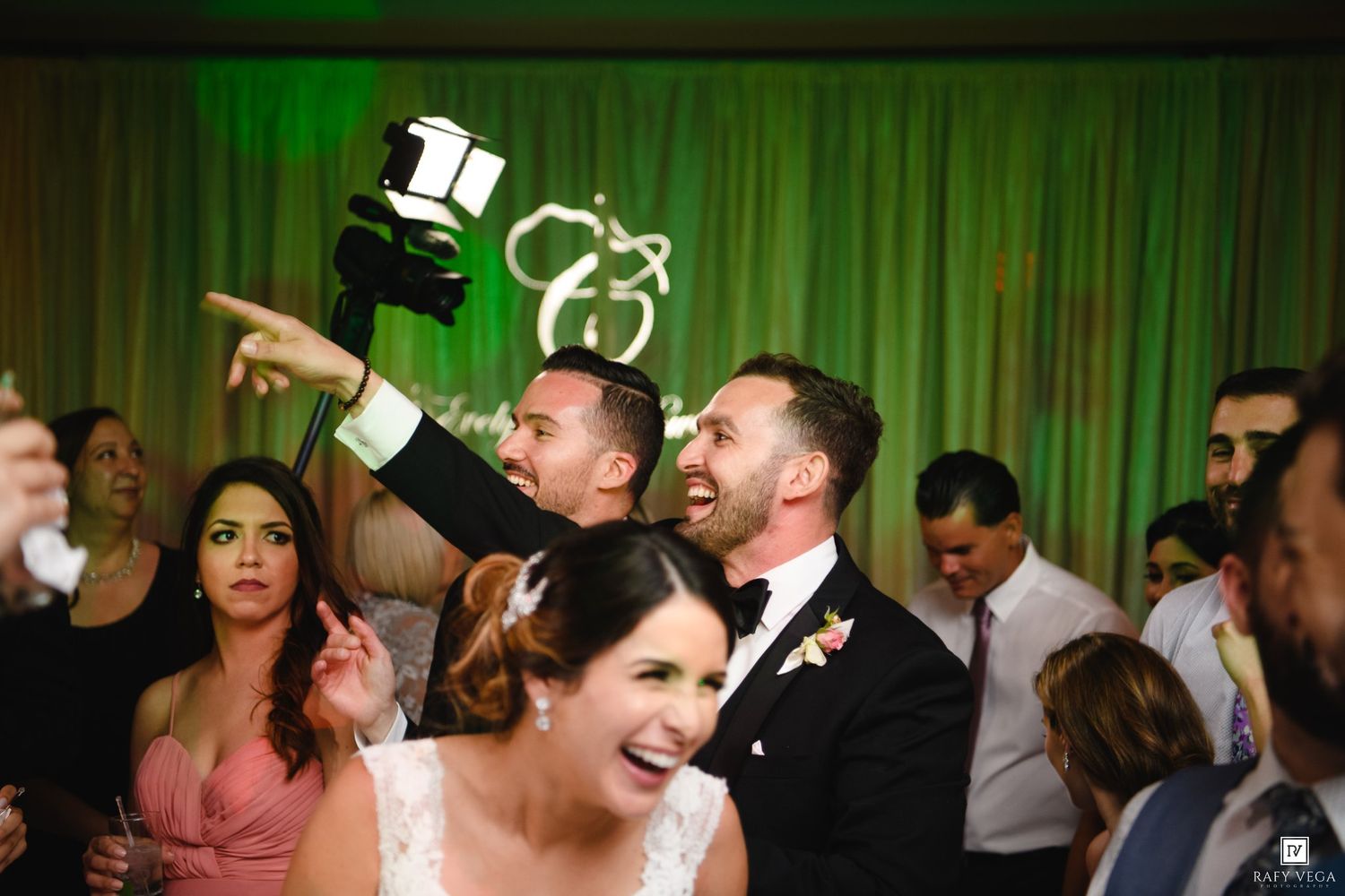 Joyful wedding reception with bride and groom celebrating, guests dancing under green lights with monogram backdrop.