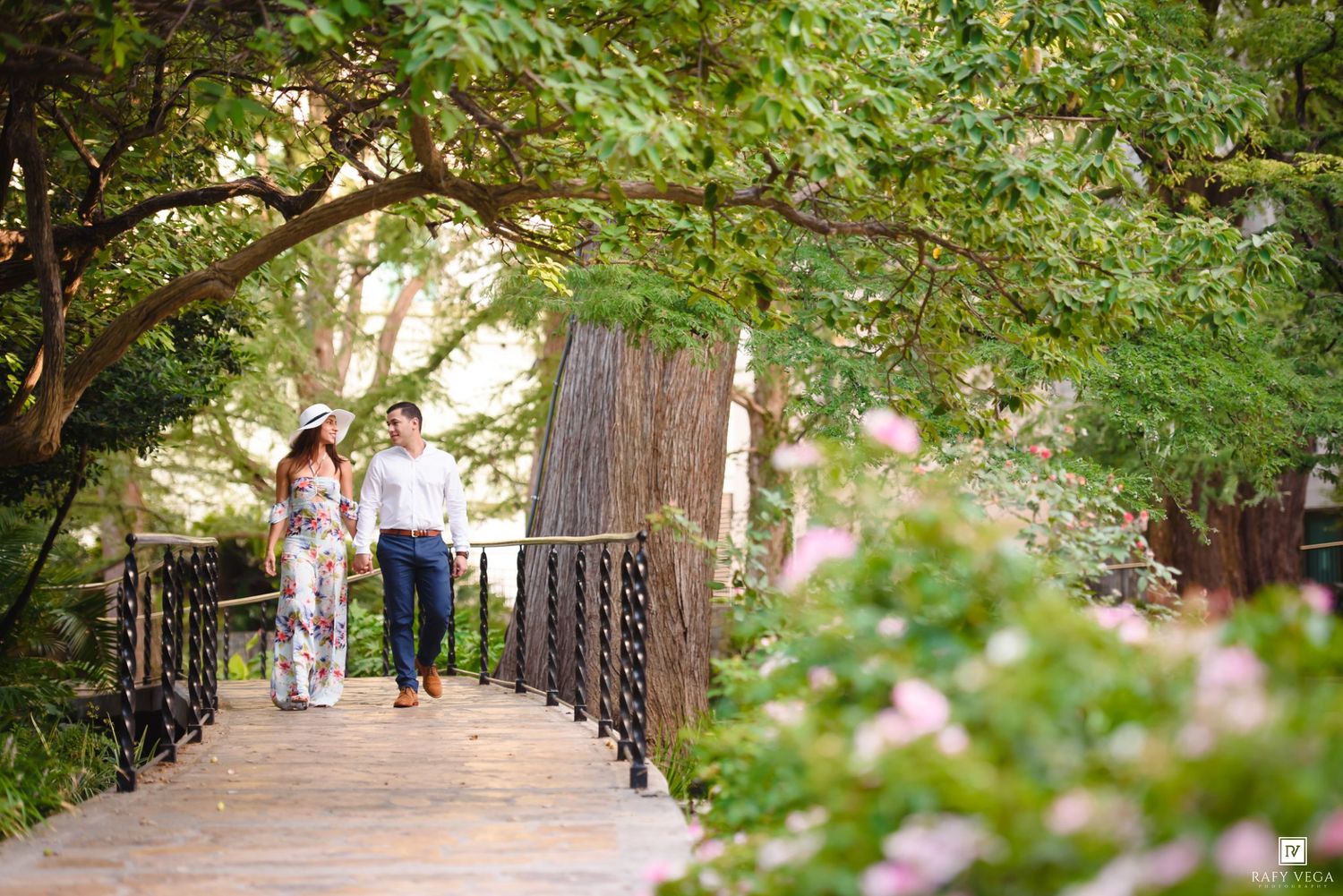 Couple walking hand-in-hand on a wooden bridge surrounded by lush green trees and pink flowers.