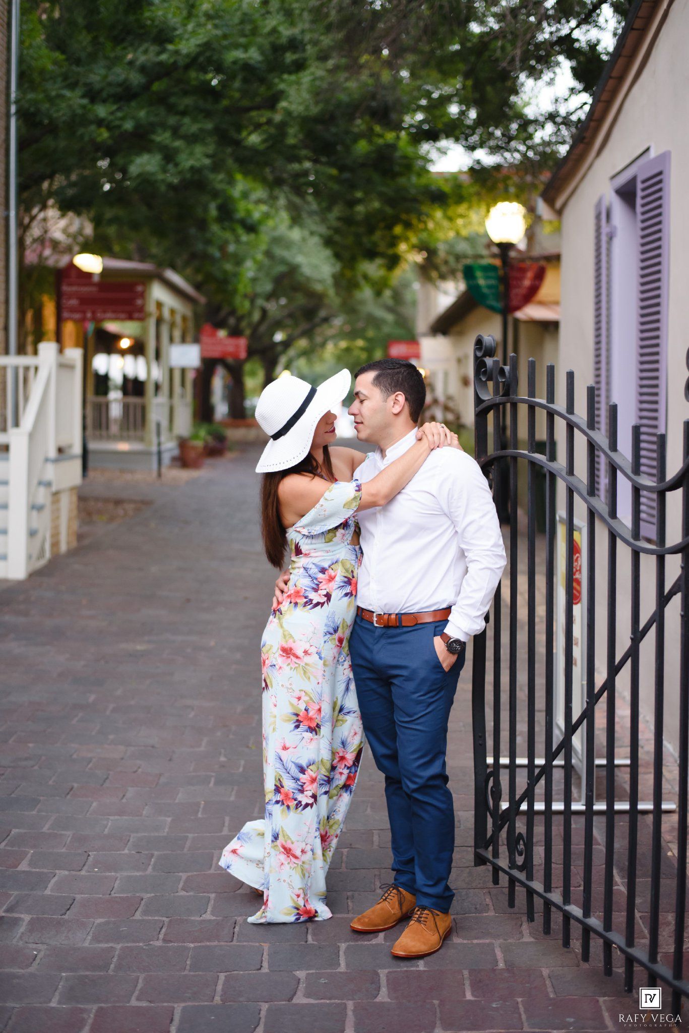 Couple sharing a romantic moment by an iron gate on a brick walkway, woman in floral dress and white hat.