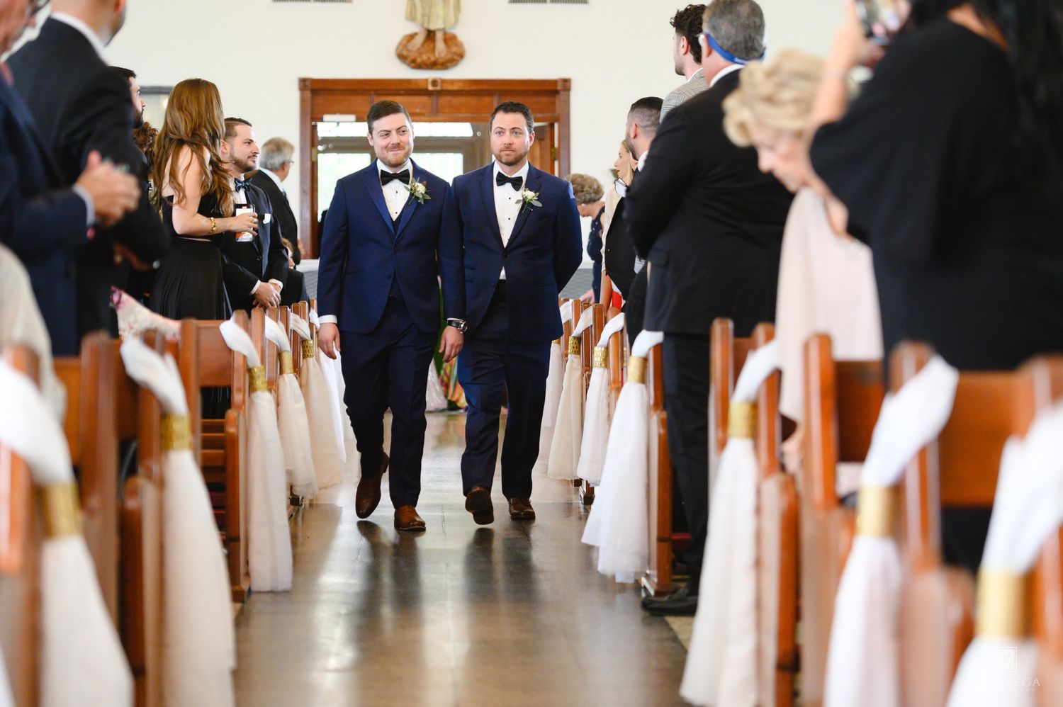 Two grooms in navy suits walk down church aisle together at elegant wedding ceremony with guests seated.