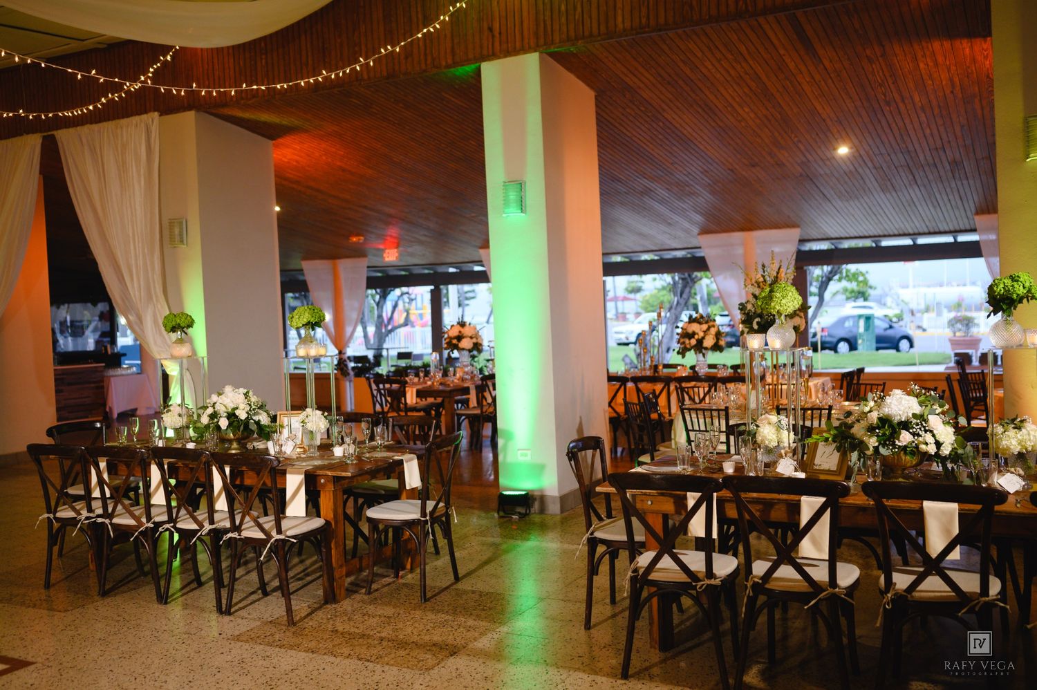 Elegant wedding reception venue with wooden cross-back chairs, floral centerpieces, string lights, and green uplighting on white columns.