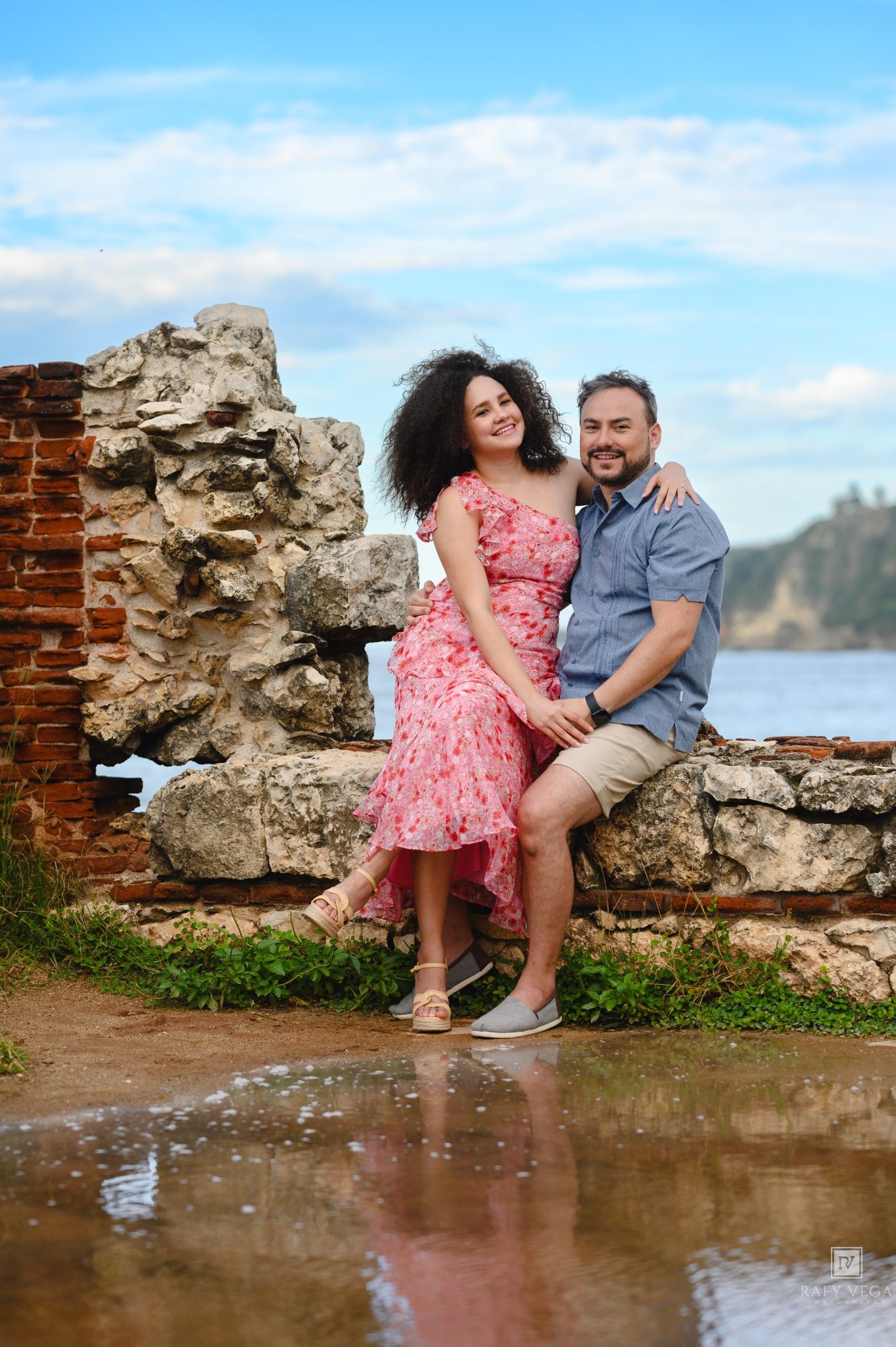 Couple smiling and embracing on stone ruins by the water, woman in pink floral dress, man in blue shirt, sunny day.