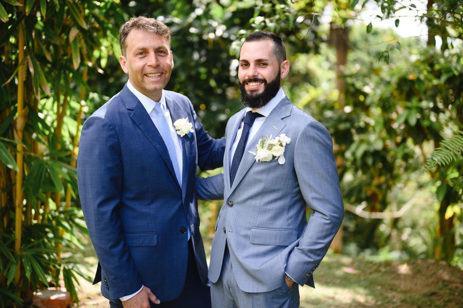 Two men in blue suits with boutonnieres posing outdoors at a wedding ceremony surrounded by greenery.