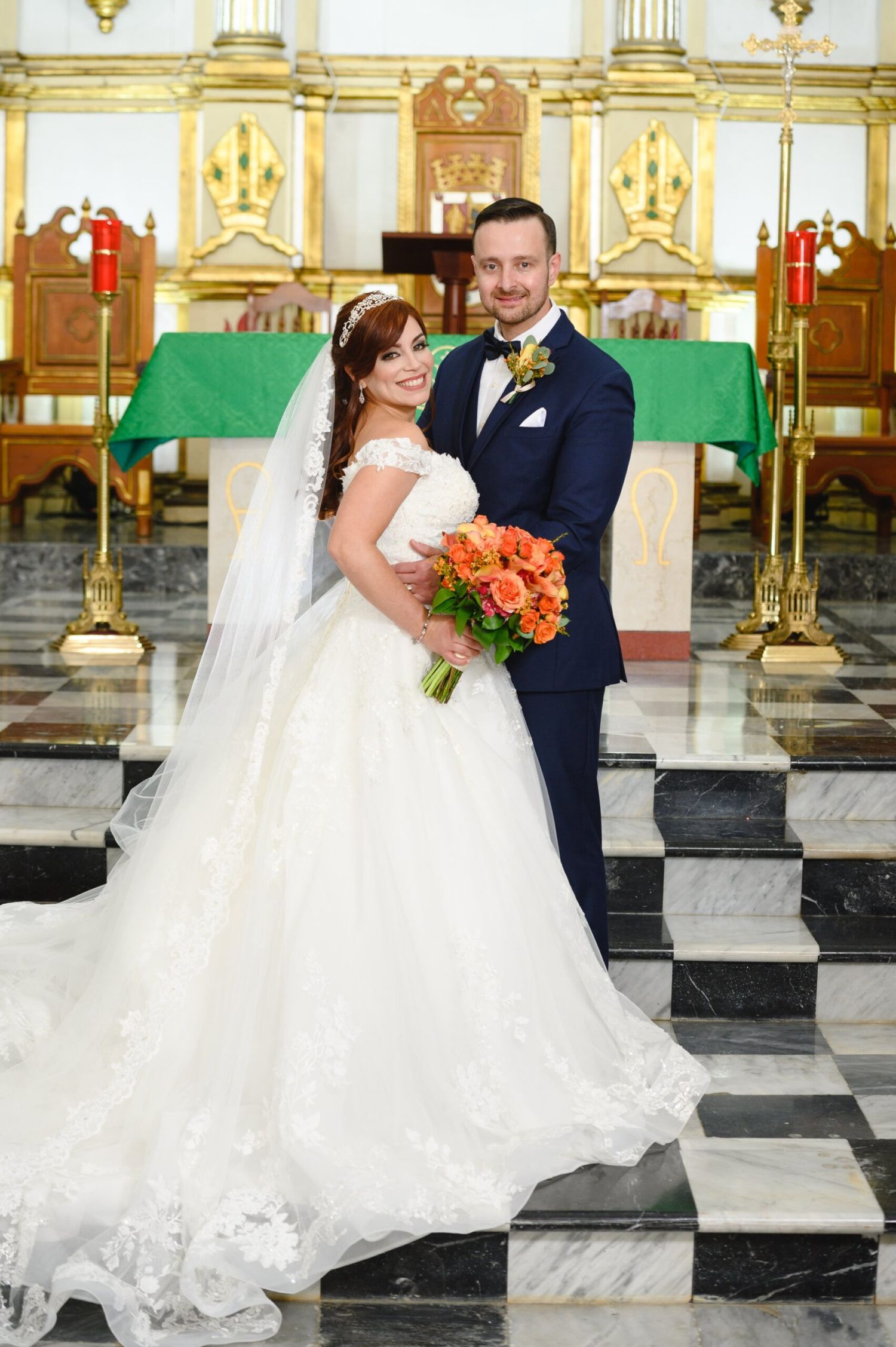 Bride in white ball gown and groom in navy suit pose together at church altar with orange bouquet.