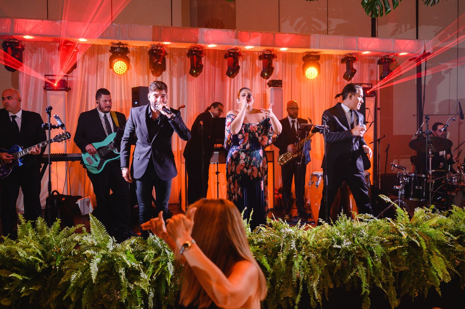 Live wedding band performing on stage with red lighting, musicians in suits playing guitars and singing at elegant reception.