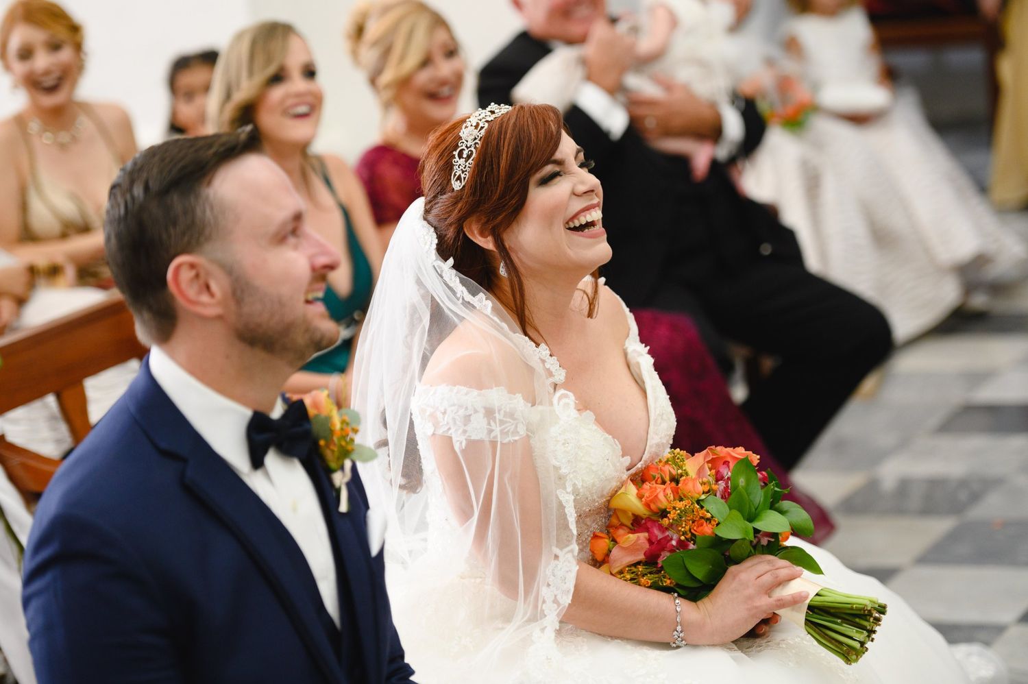 Laughing bride in white gown with tiara and groom in navy suit during joyful wedding ceremony.