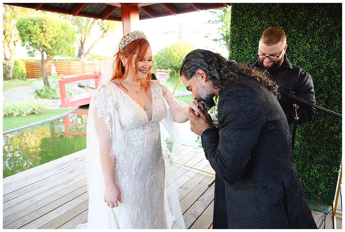 Groom kisses bride's hand tenderly during outdoor wedding ceremony in garden setting.