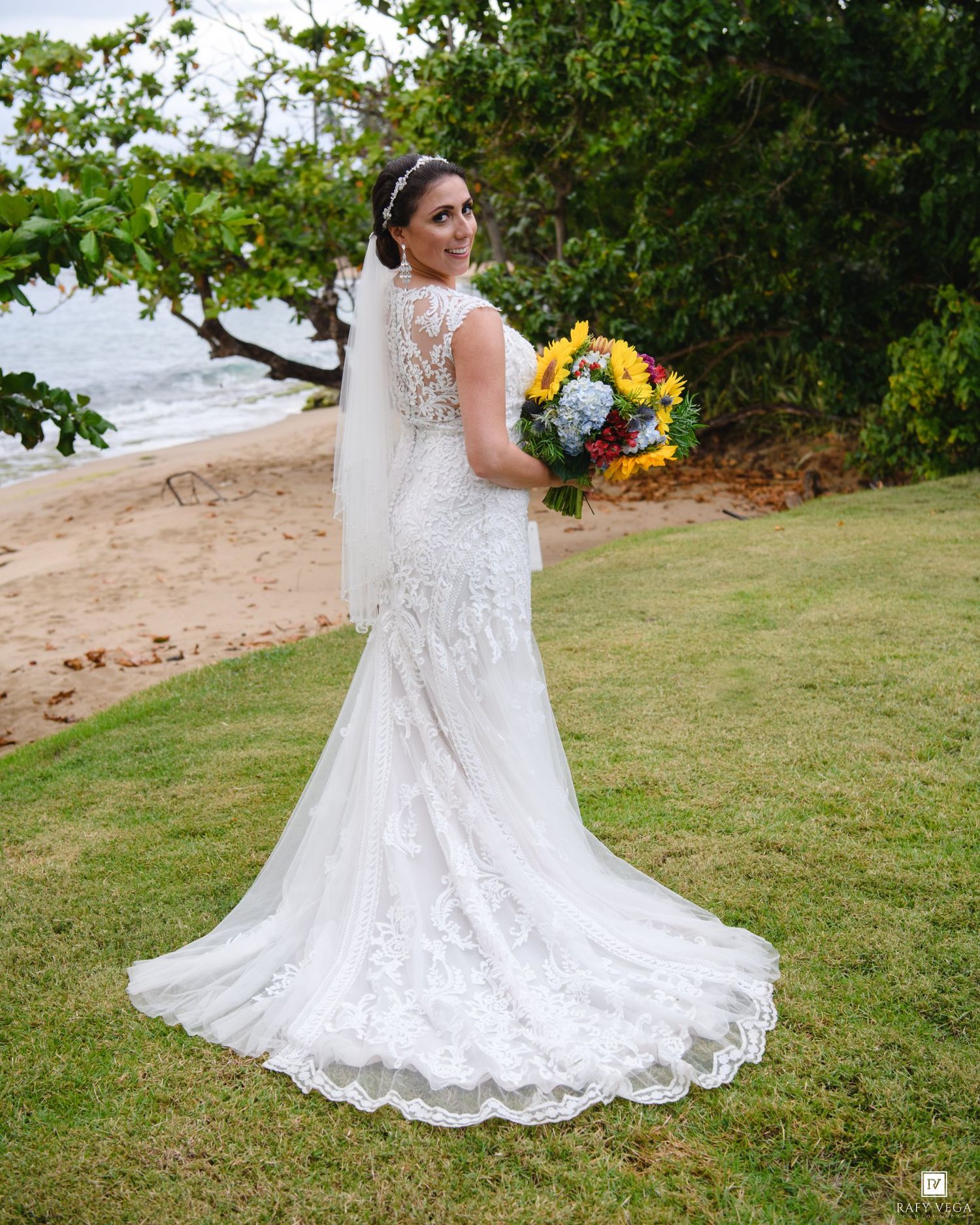 Bride in lace mermaid wedding gown with veil holding colorful bouquet on tropical beach lawn.