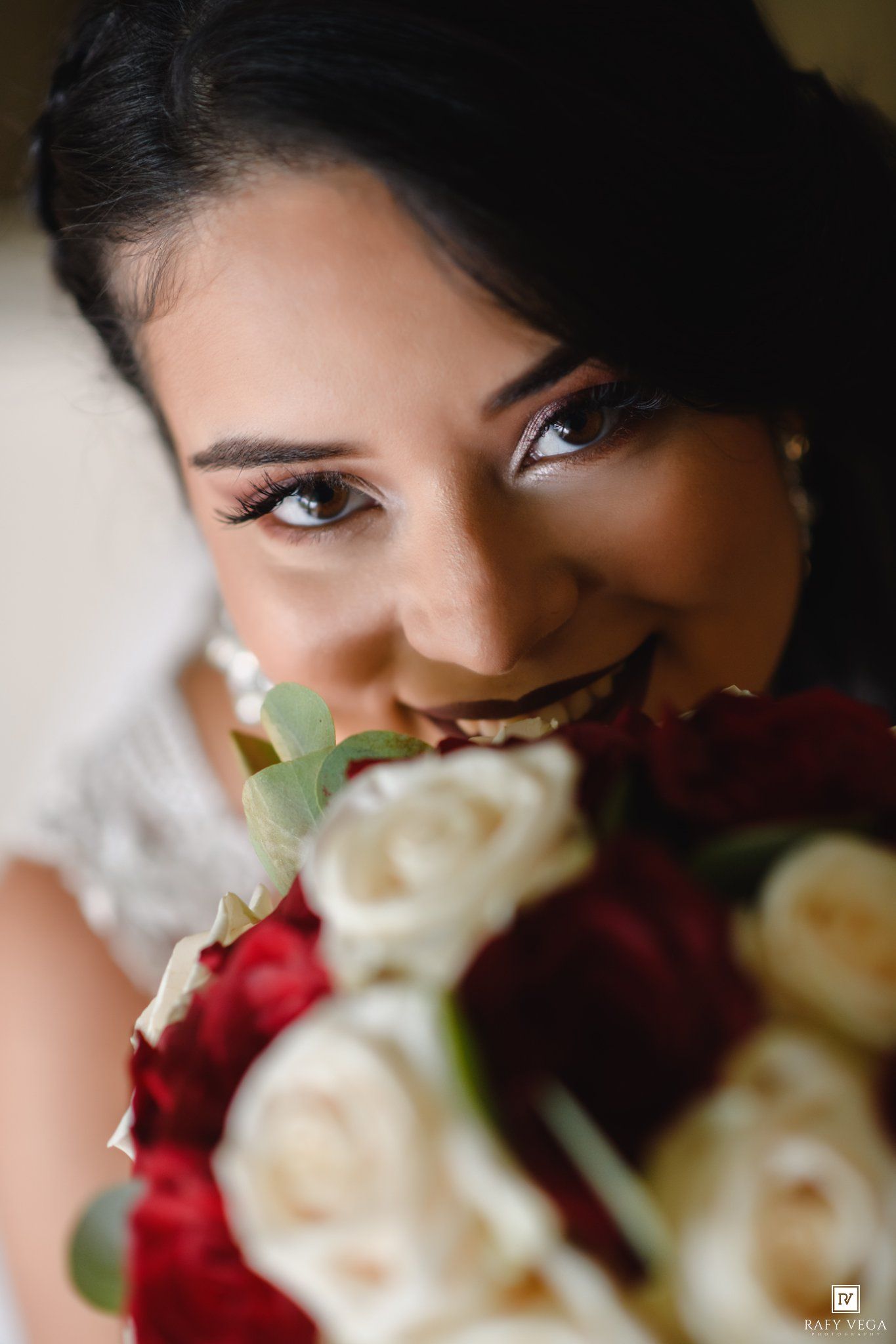 Smiling bride holding red and white rose bouquet close to her face, showcasing elegant makeup and dark hair.