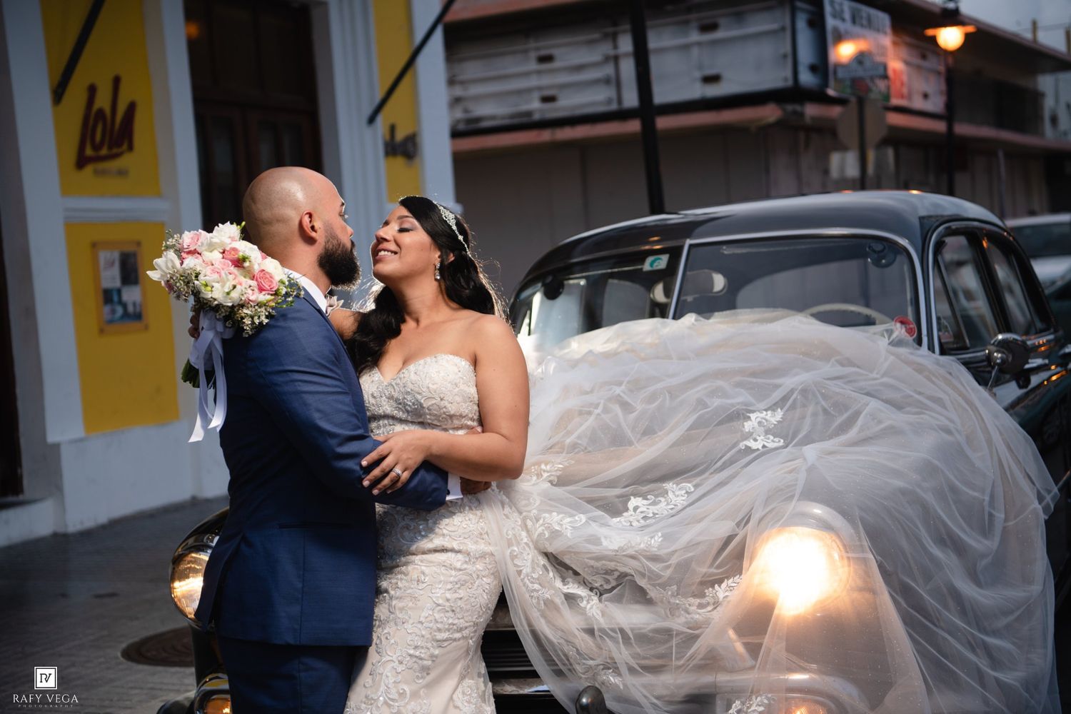 Bride and groom share romantic kiss beside vintage black car on cobblestone street at dusk.