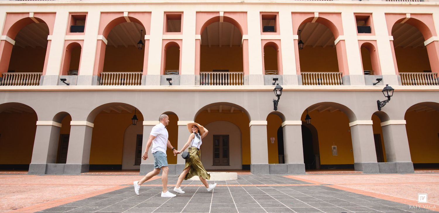 Two people walking through a sunlit colonial courtyard with arched arcades and terracotta facade.