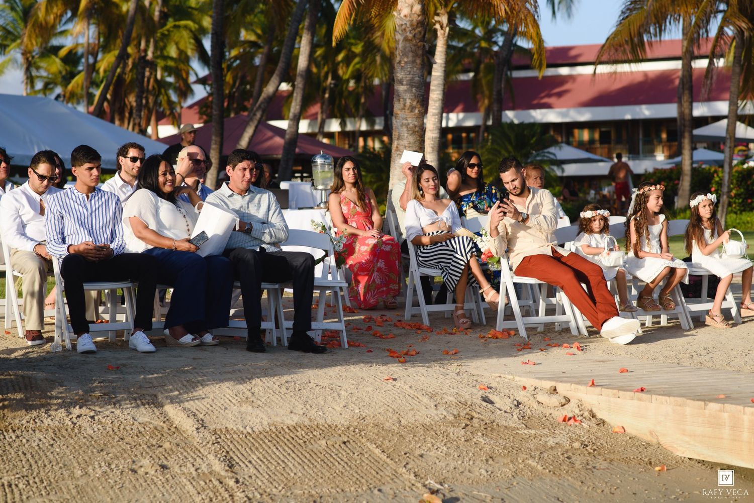Guests seated on white chairs at an outdoor beach wedding ceremony with palm trees and a tropical resort in the background.