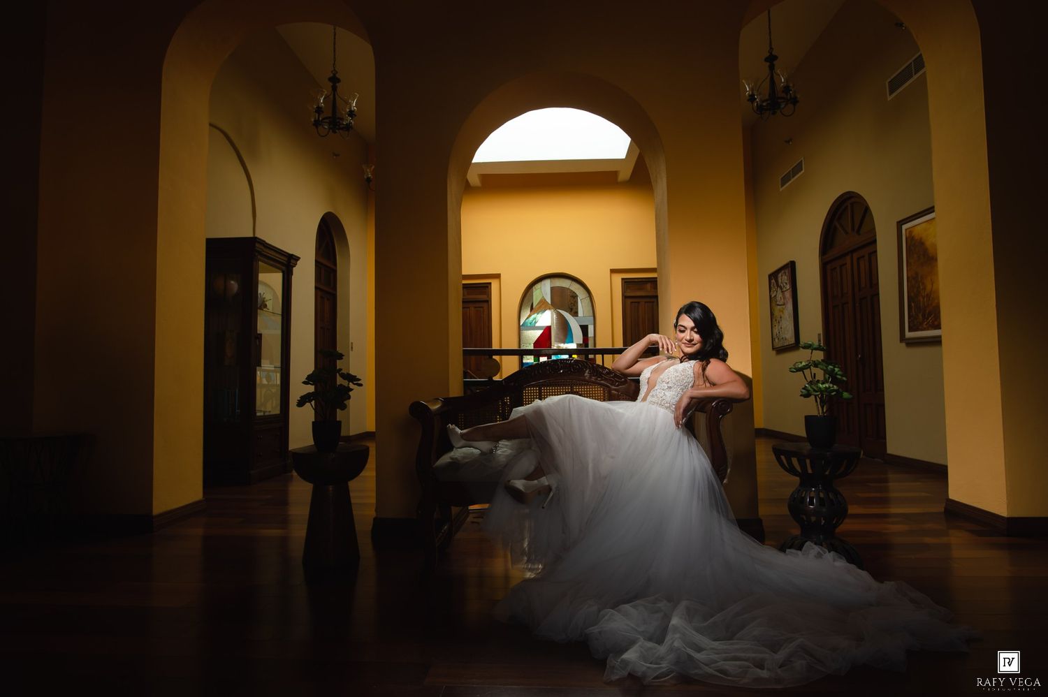 Bride in flowing white wedding dress posed gracefully in a golden-lit vintage corridor with arched architecture.