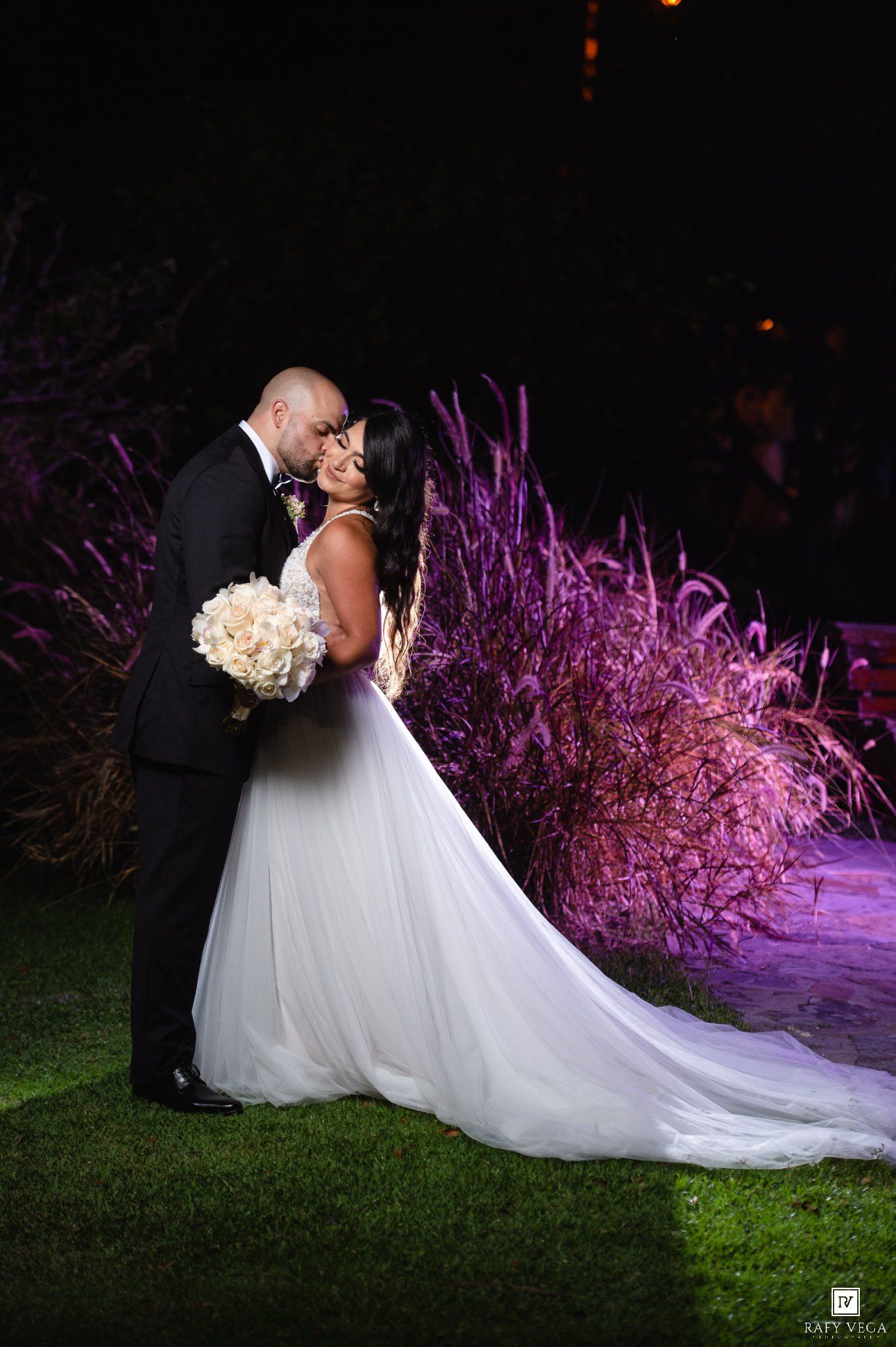 Bride and groom share a romantic kiss at night, surrounded by purple-lit ornamental grasses in a lush garden setting.
