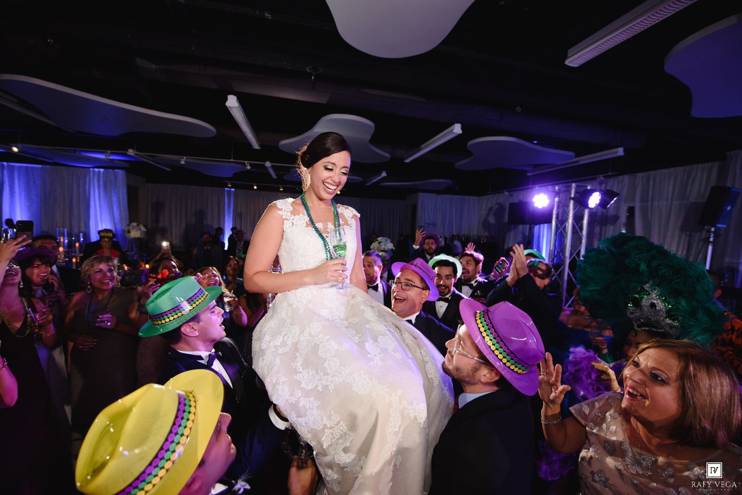 Bride lifted by guests in colorful hats during lively wedding reception celebration.