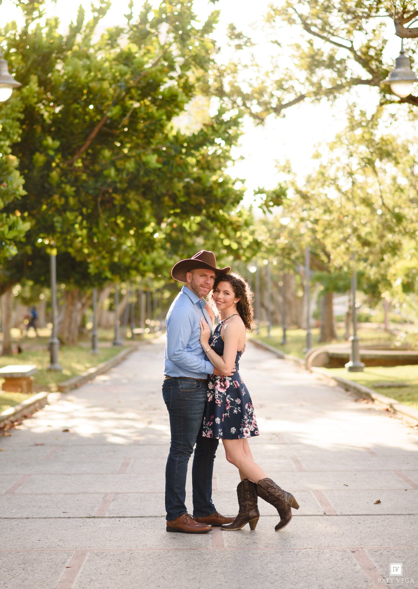 Couple posing on a tree-lined path, man in cowboy hat and woman in floral dress with boots.