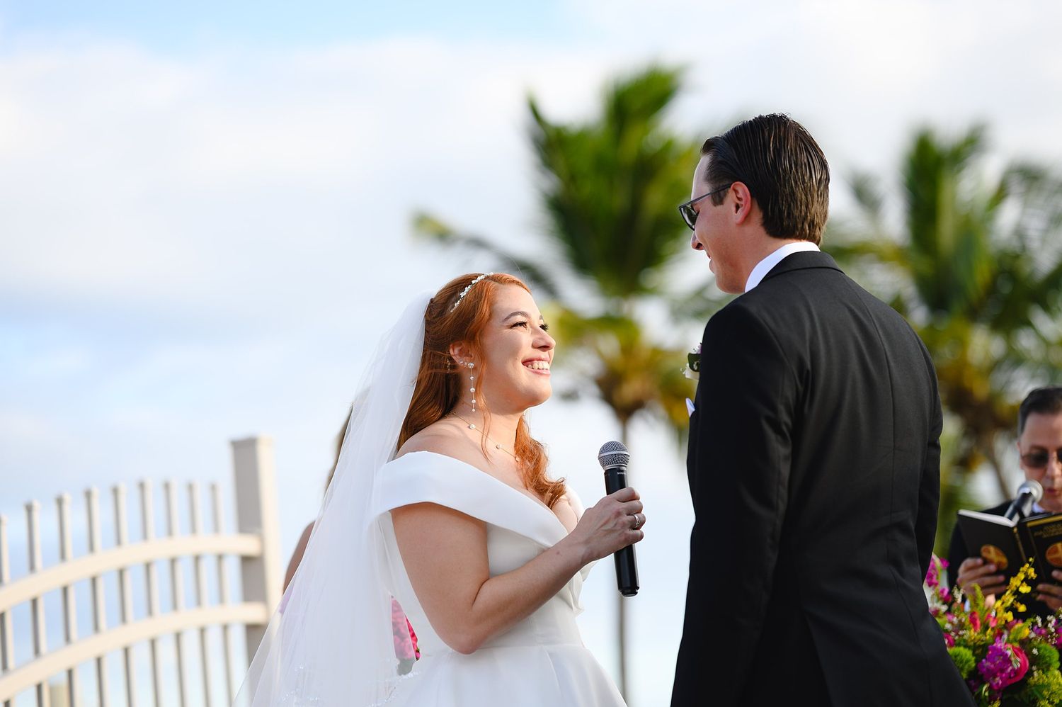 Smiling bride in white gown and veil exchanges vows with groom at outdoor beach wedding ceremony.
