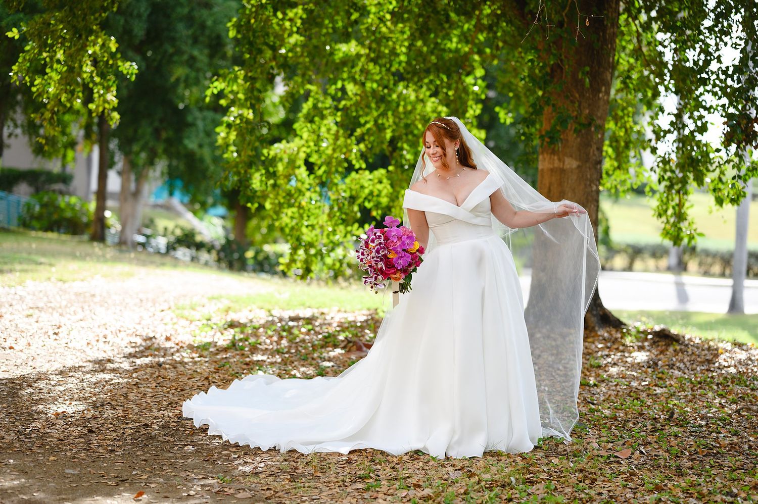 Bride in white off-shoulder gown with long train holds pink bouquet under green tree outdoors.