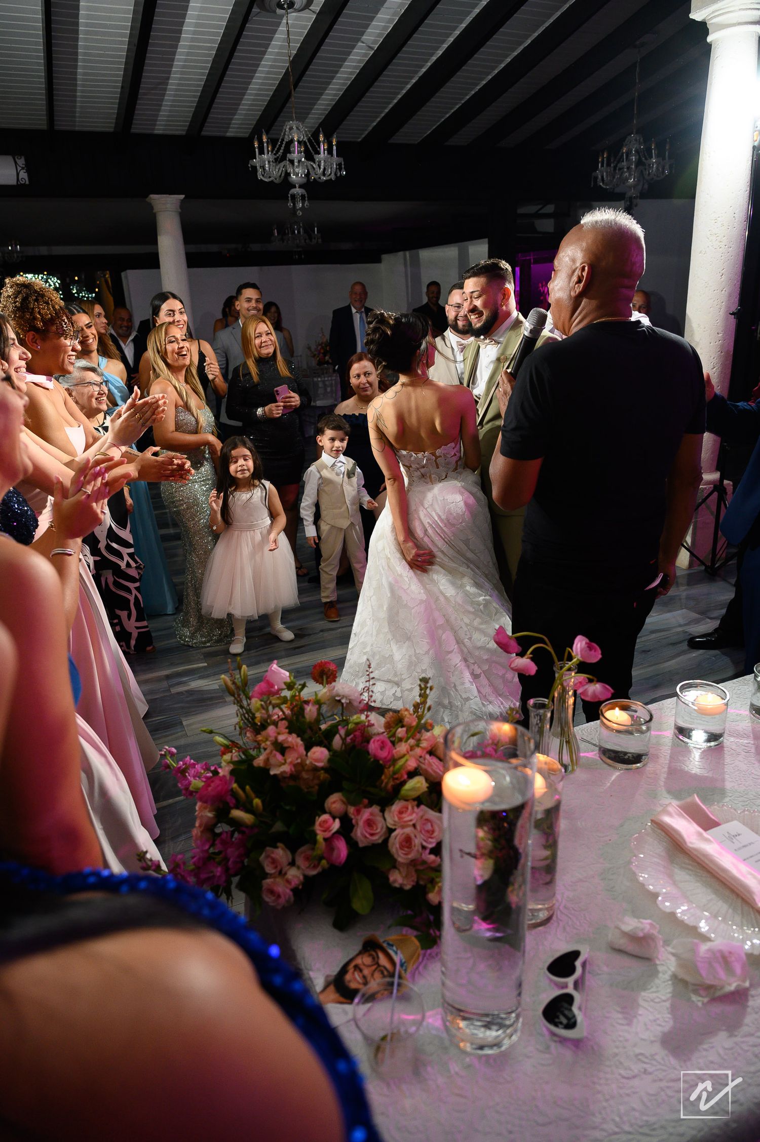 Bride in a lace wedding gown stands before smiling guests as a man speaks into a microphone near a candlelit table with pink flowers.