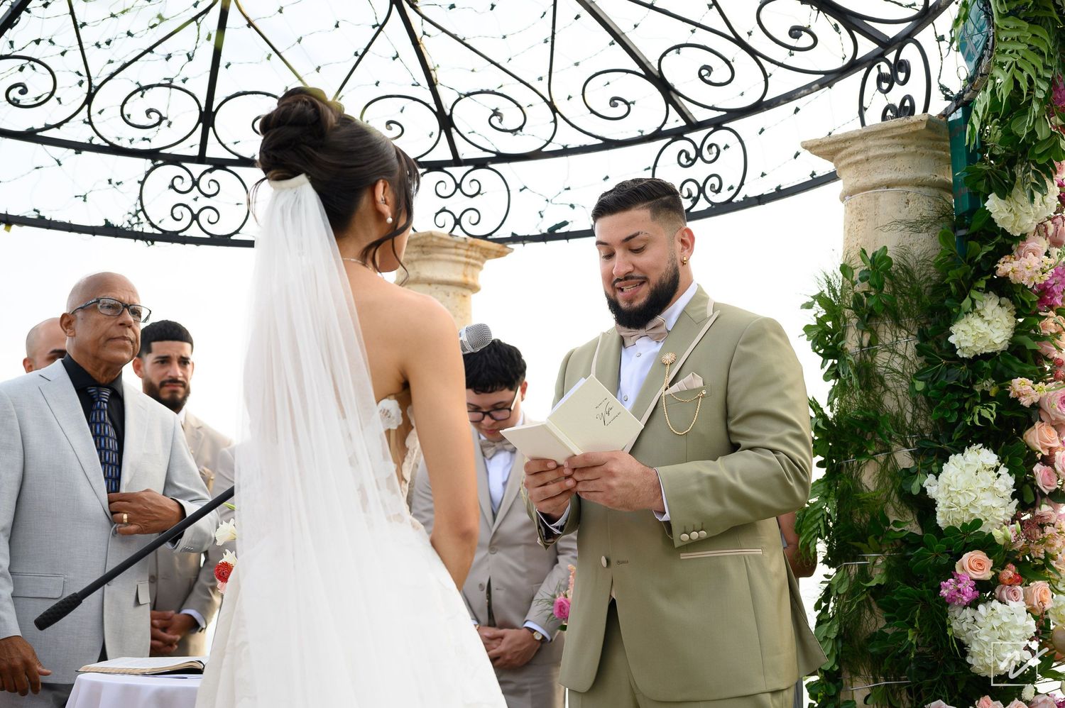 Bride in a white gown and veil faces a bearded groom in a tan suit reading vows during an outdoor wedding ceremony beside a floral arch.